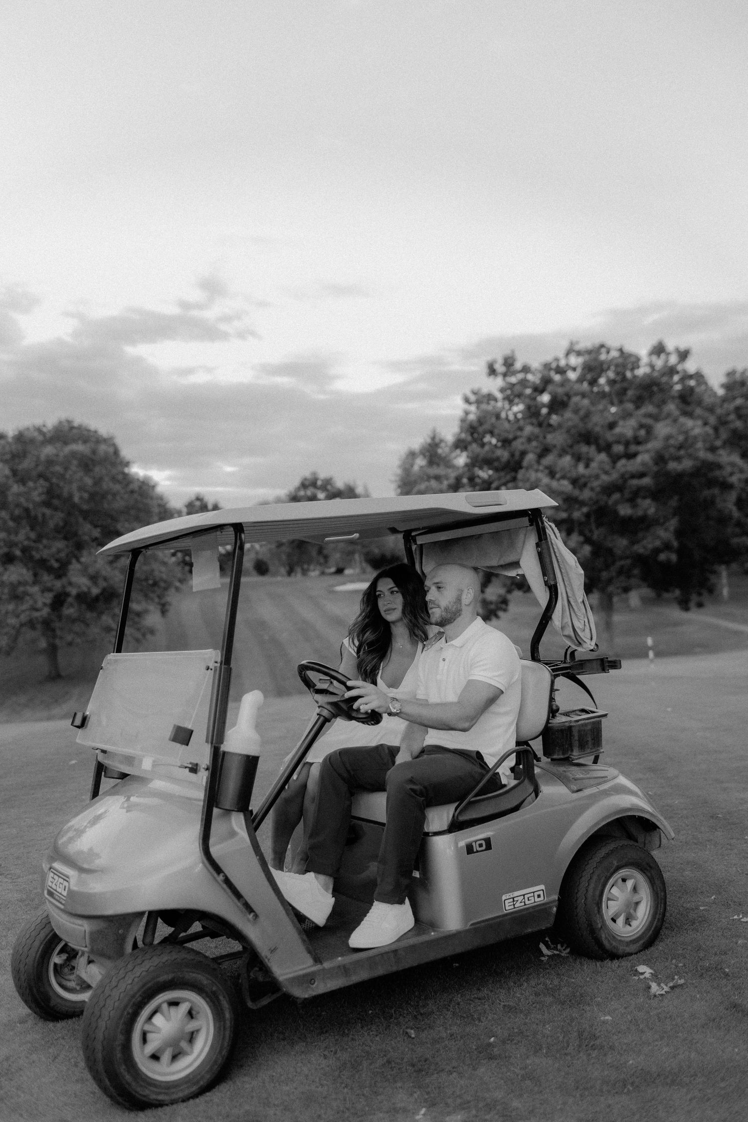 Black and white image of couple driving golf cart on course at dusk.