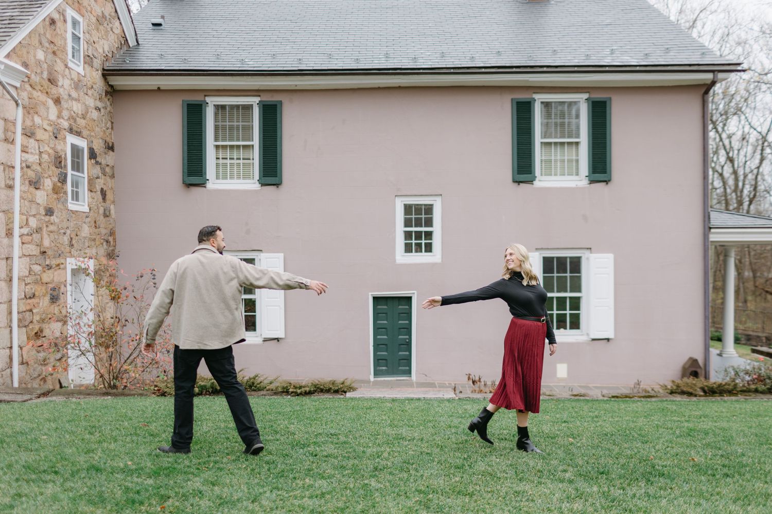 A playful couple dances in front of a pink house with green shutters.