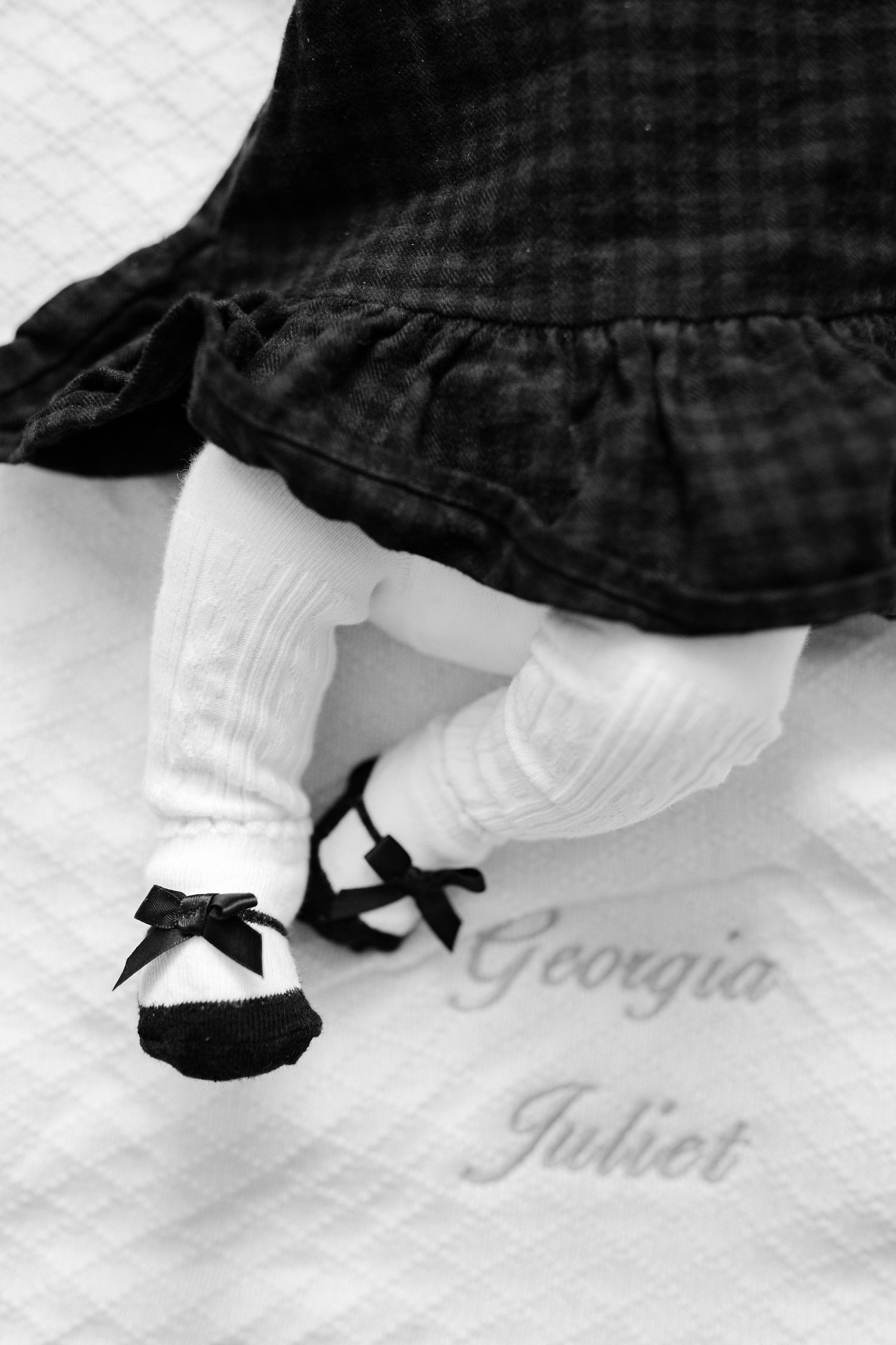 Black and white photo of baby feet wearing white socks with black bow shoes.