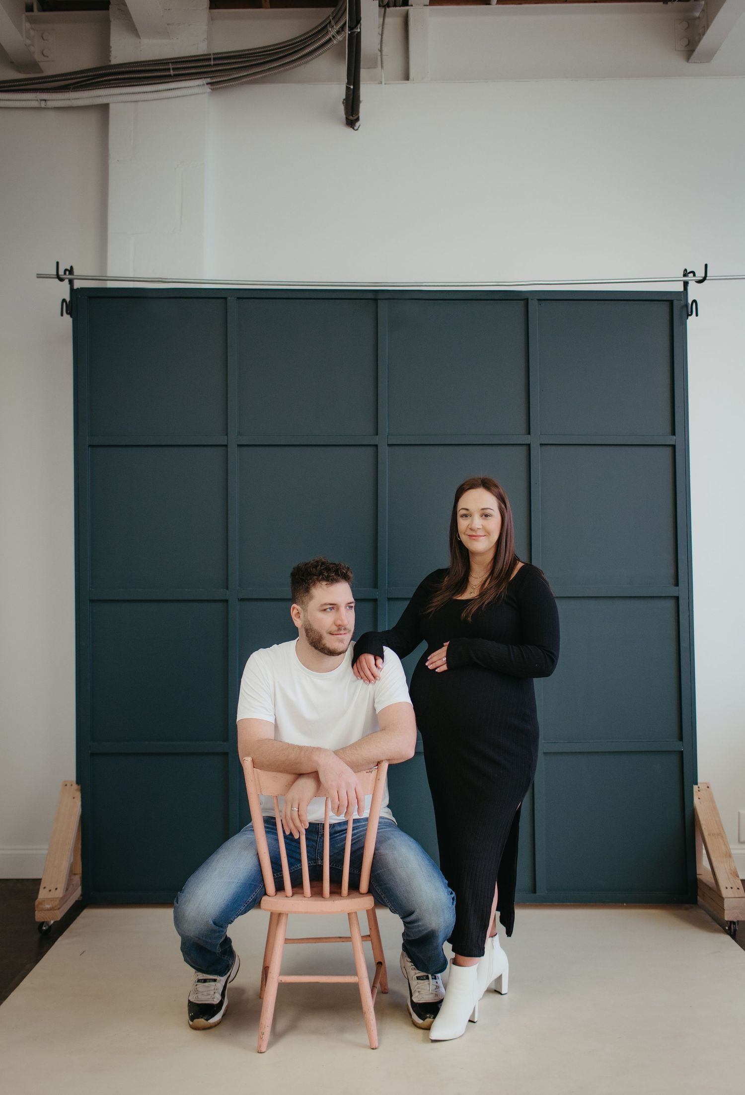Modern maternity photoshoot with a teal backdrop and wooden chair.