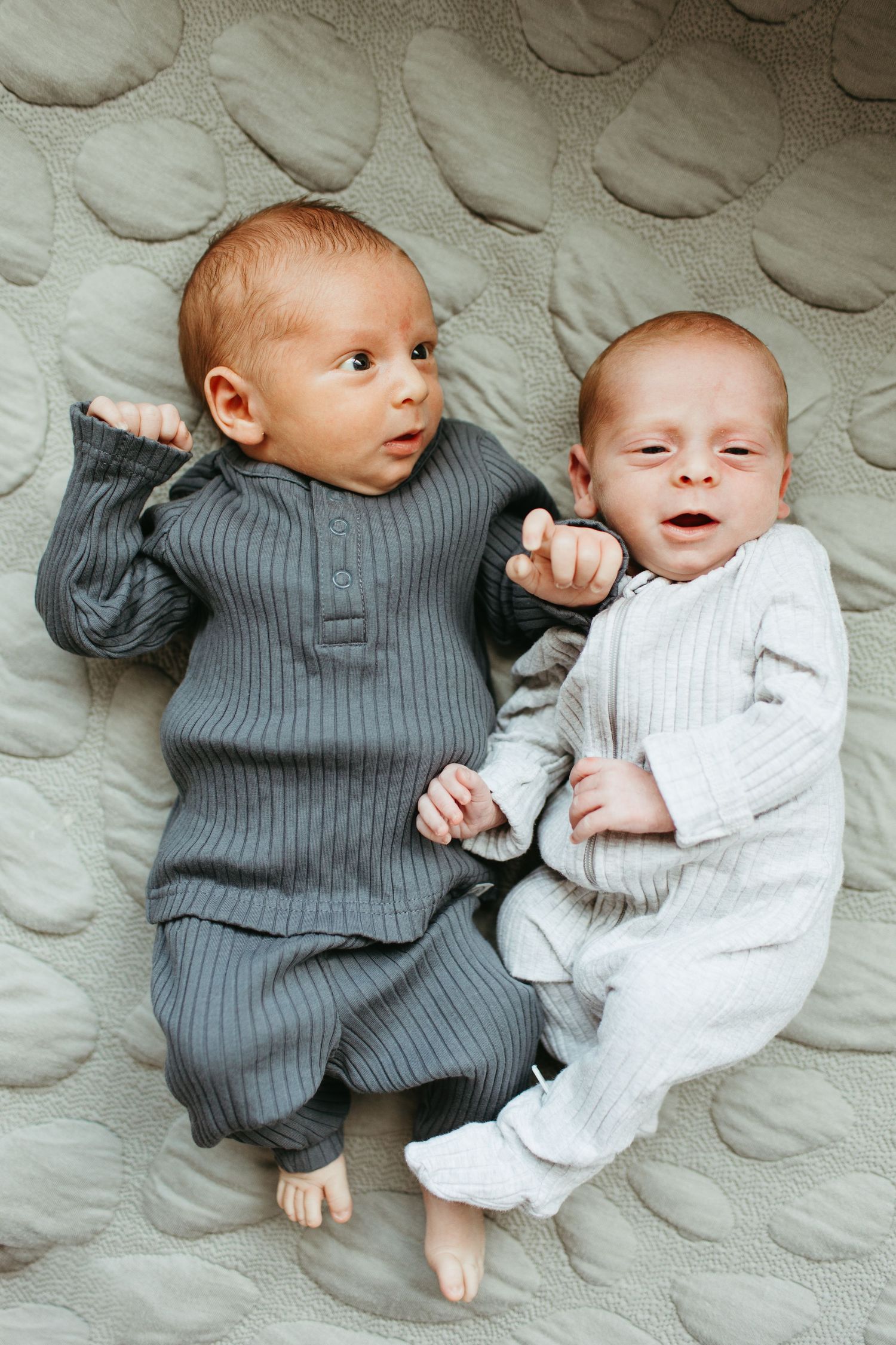 Two babies in knit outfits, one gray and one white, lie together on a textured blanket.