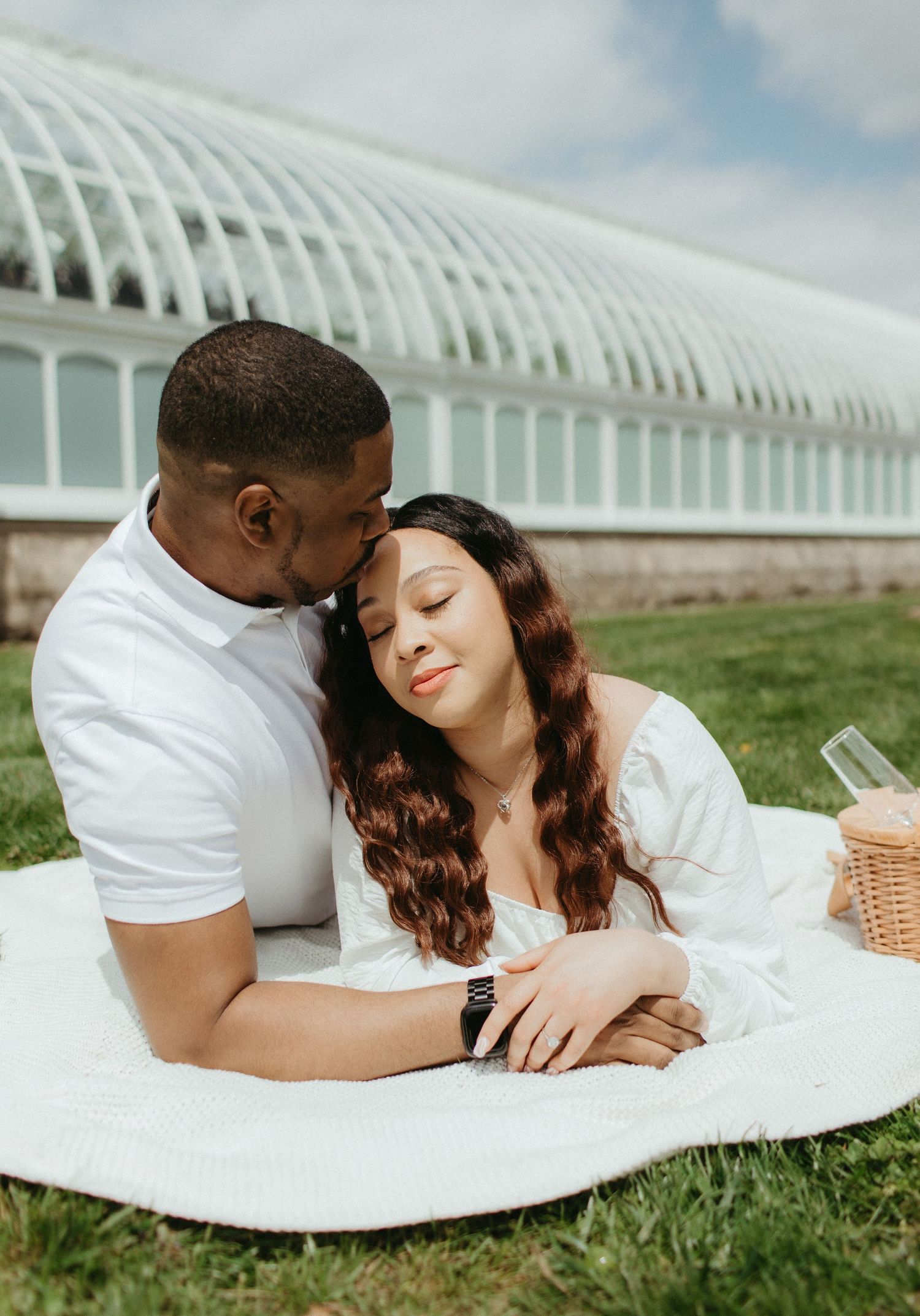 A couple enjoys an intimate picnic moment on a blanket near a greenhouse structure.