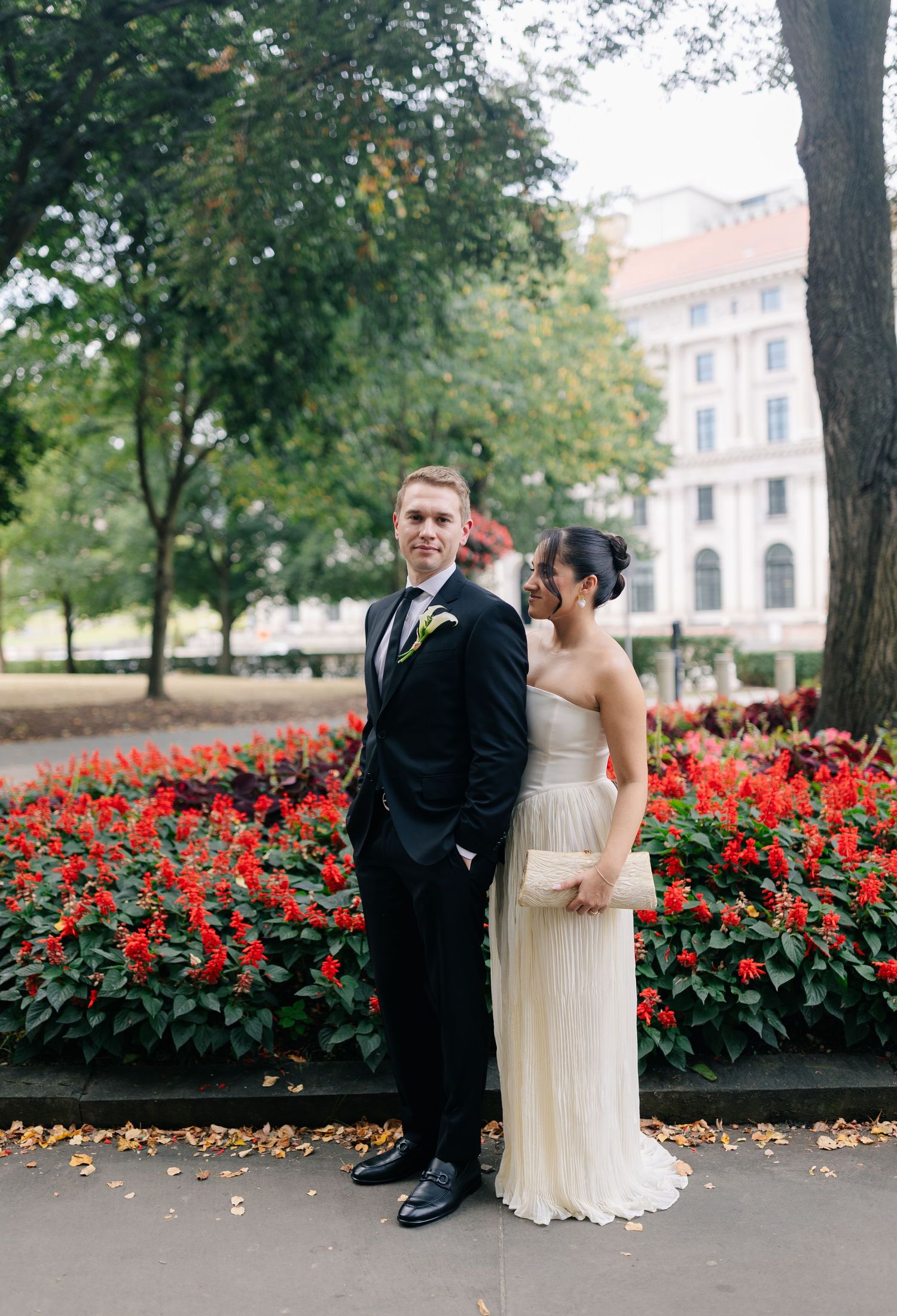 A couple poses in front of vibrant red flowers and trees during an autumn wedding photoshoot.