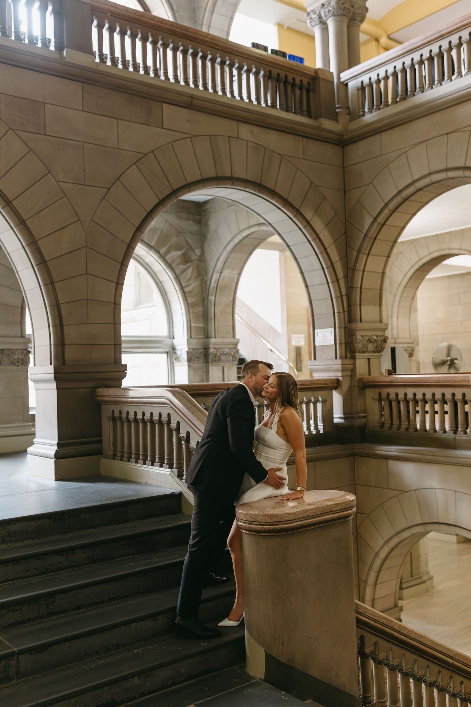A couple shares a romantic moment in an elegant stone archway interior with ornate balustrades and classical architecture.