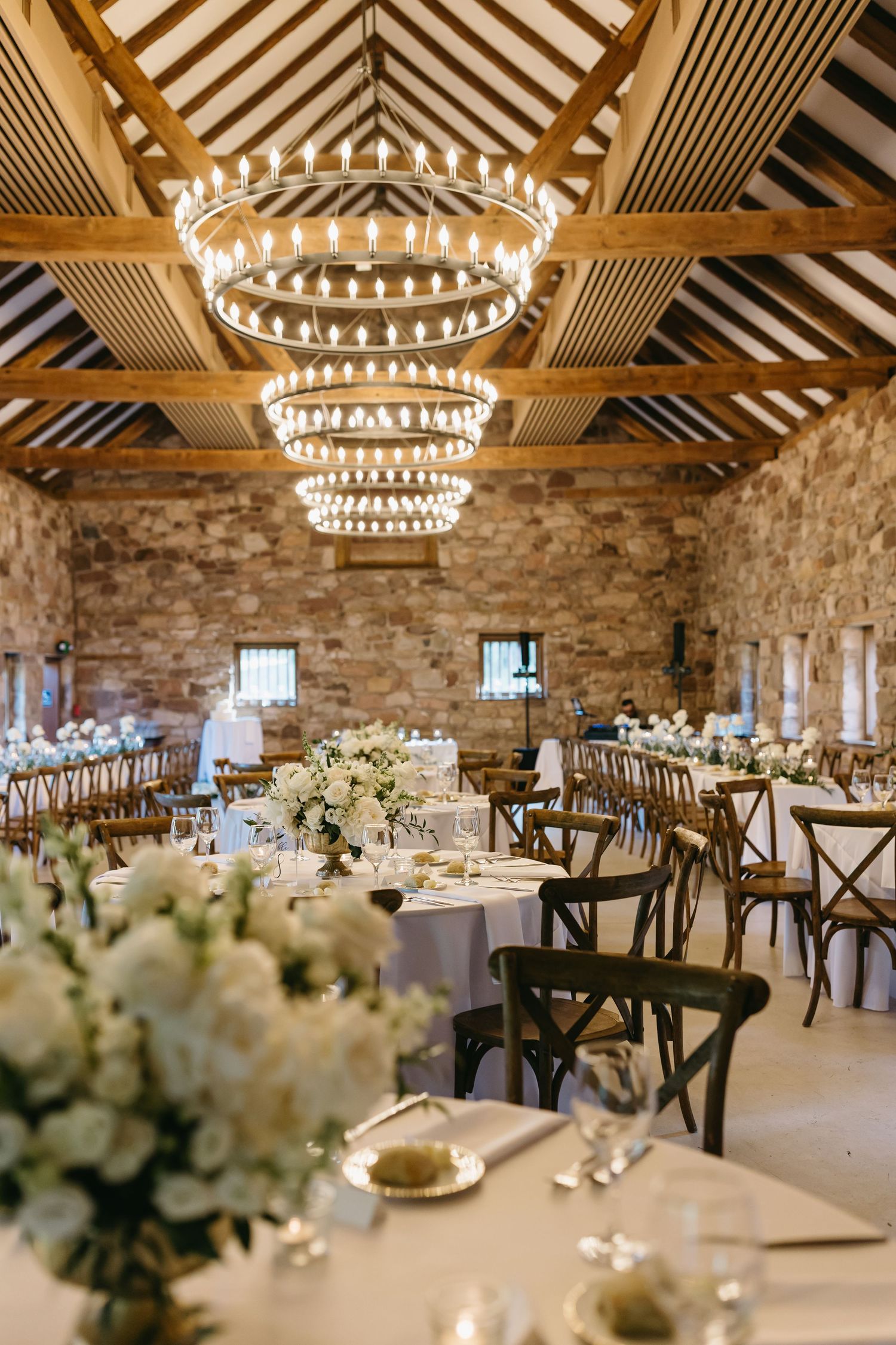 Elegant rustic wedding reception venue with tiered chandelier, stone walls, and white floral centerpieces on round tables.
