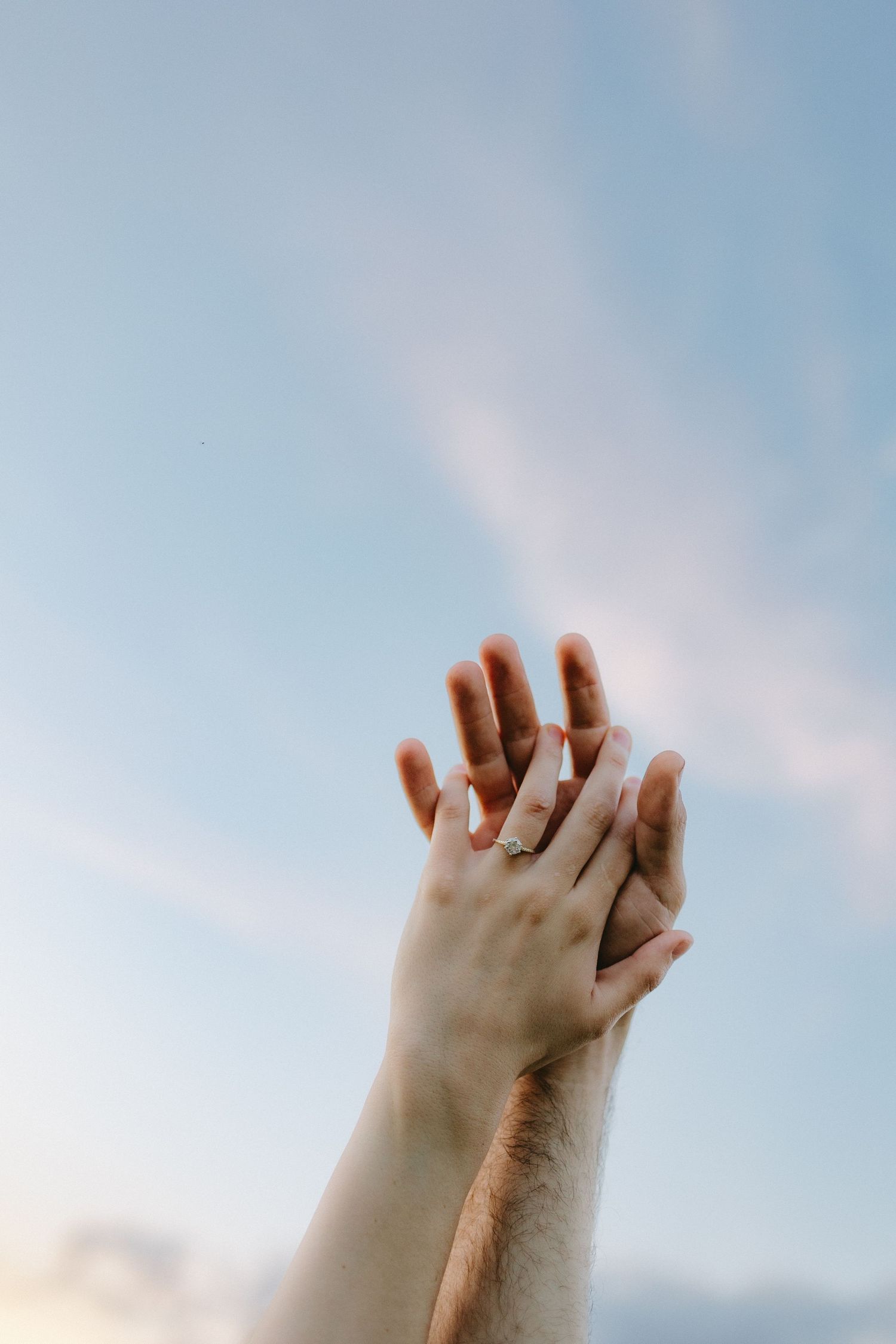 Two hands reaching towards a light blue sky with a wedding ring visible.