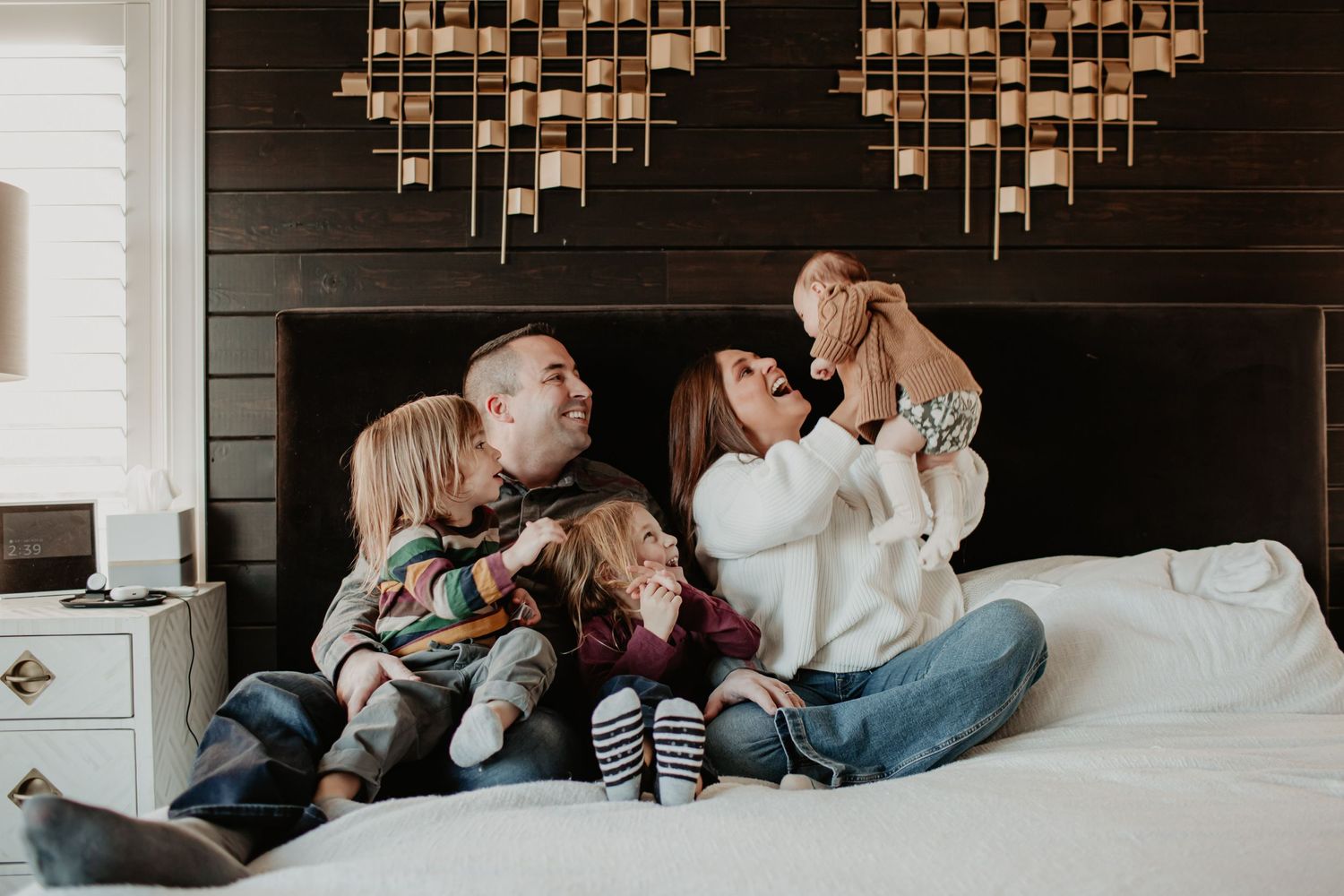 A family shares a playful moment together on a bed with decorative wall art.
