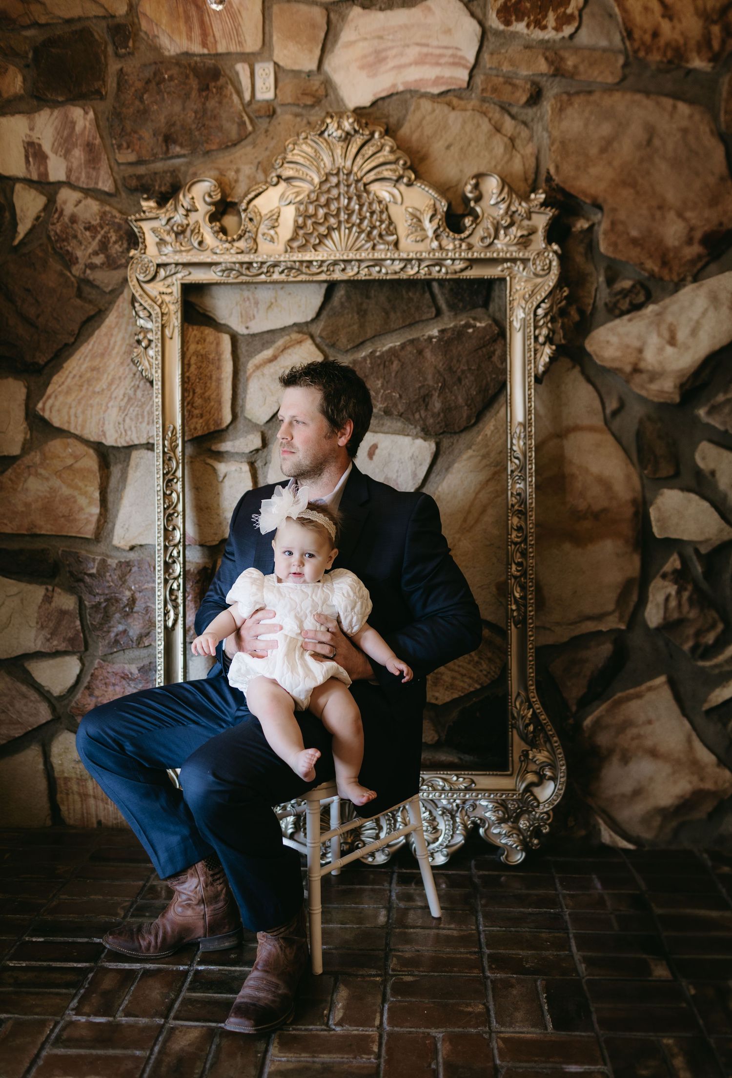 Baby and adult sit in vintage gold frame against rustic stone wall backdrop.