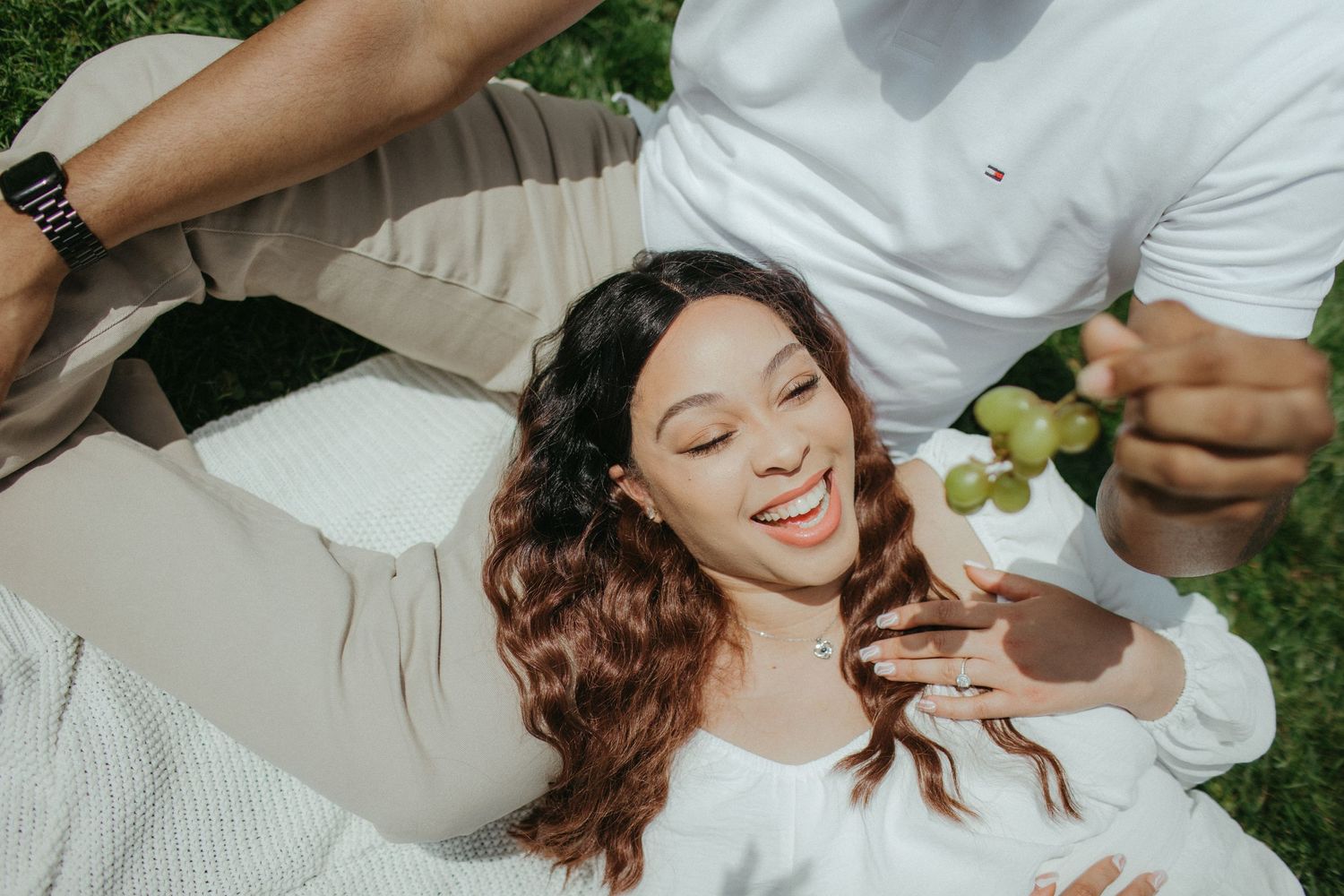 A couple enjoys a sunny picnic with fresh grapes on a white blanket outdoors.