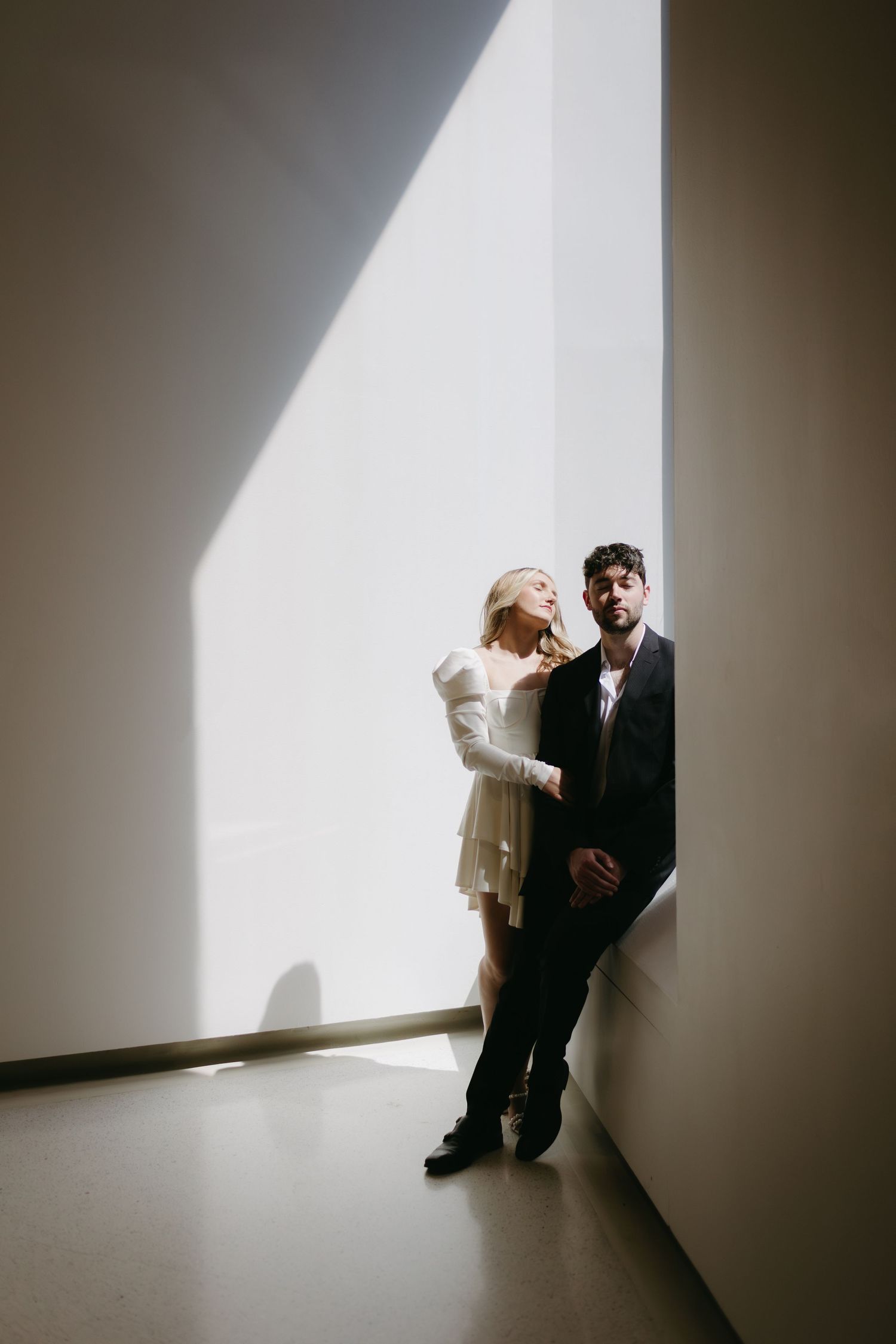Dramatic lighting creates shadow play as couple stands against minimal white wall.