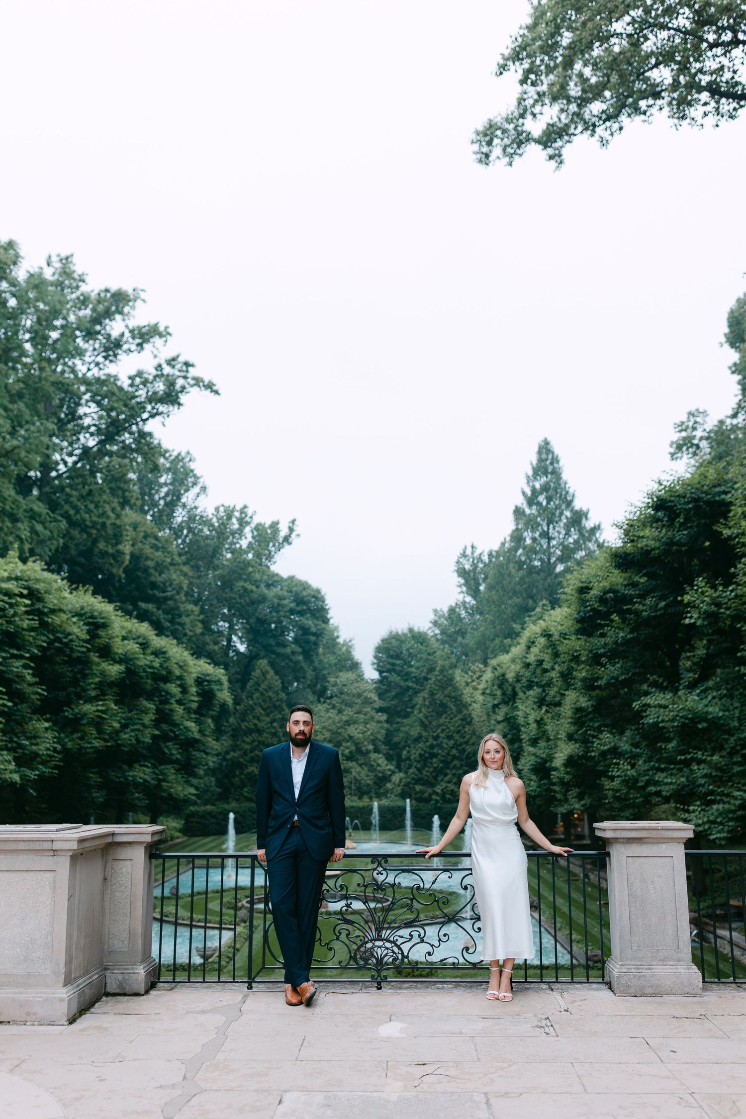 Couple posing at ornate iron gate in lush garden, man in navy suit, woman in white dress, fountain backdrop.