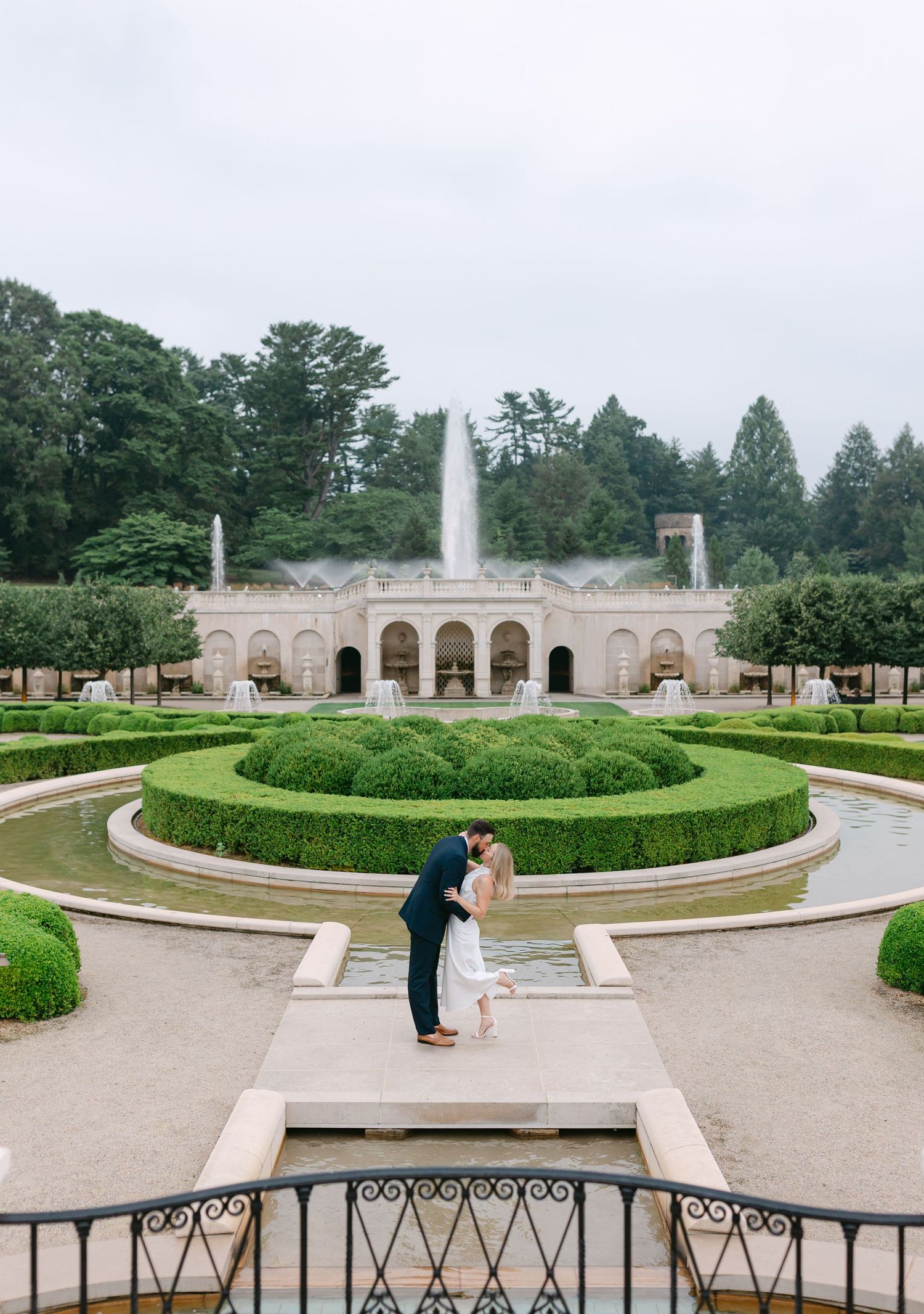 Couple sharing a romantic kiss in formal garden with fountain, manicured hedges, and elegant stone building backdrop.