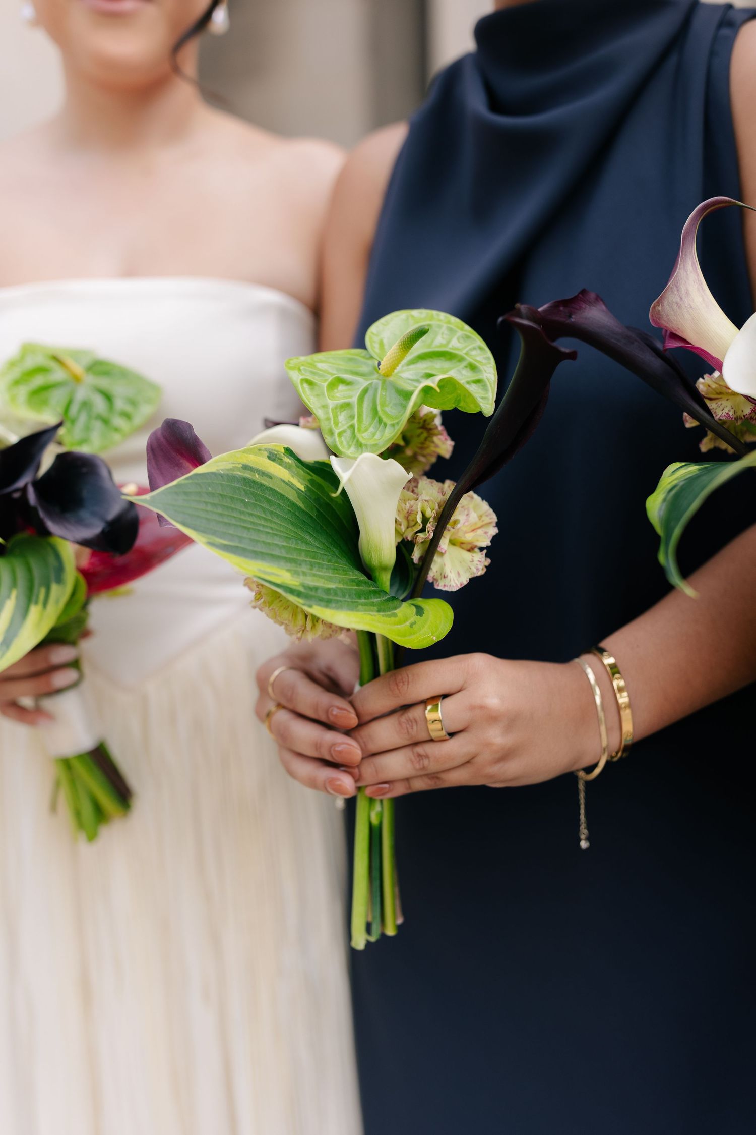 Wedding party members hold bouquets with white calla lilies and green tropical leaves against navy dresses.
