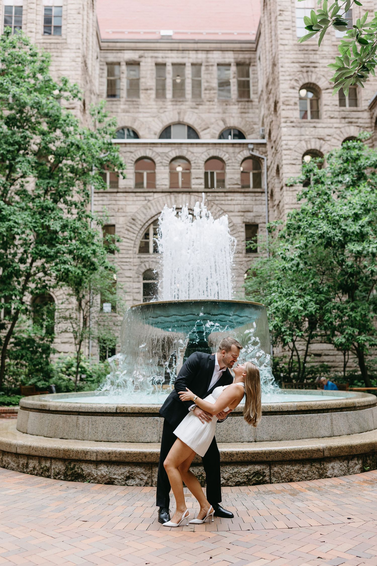 Romantic couple embracing in front of ornate stone fountain at historical building courtyard with arches and greenery.