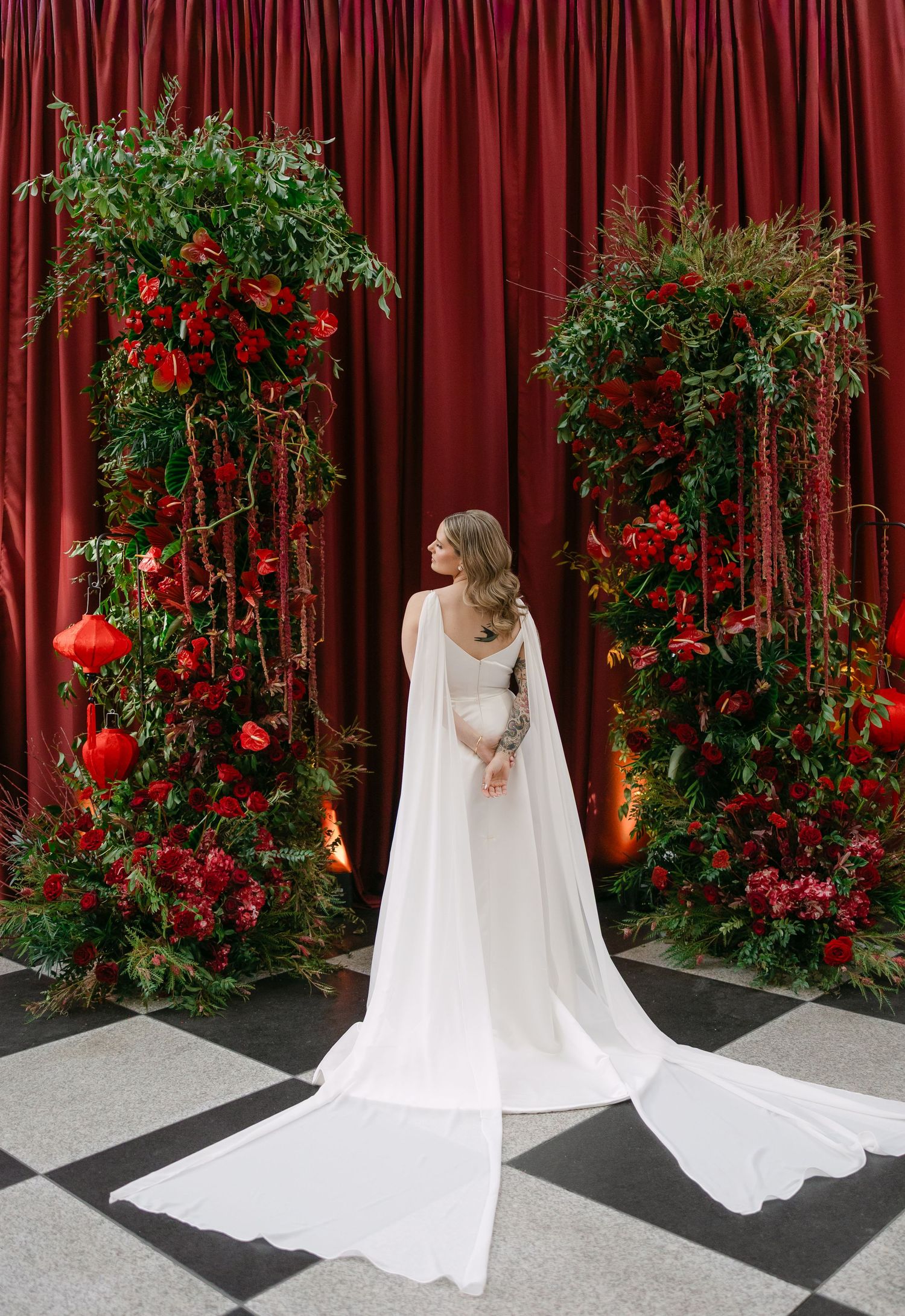 A flowing white wedding dress and veil against a red curtain backdrop with climbing roses creates an elegant bridal scene.