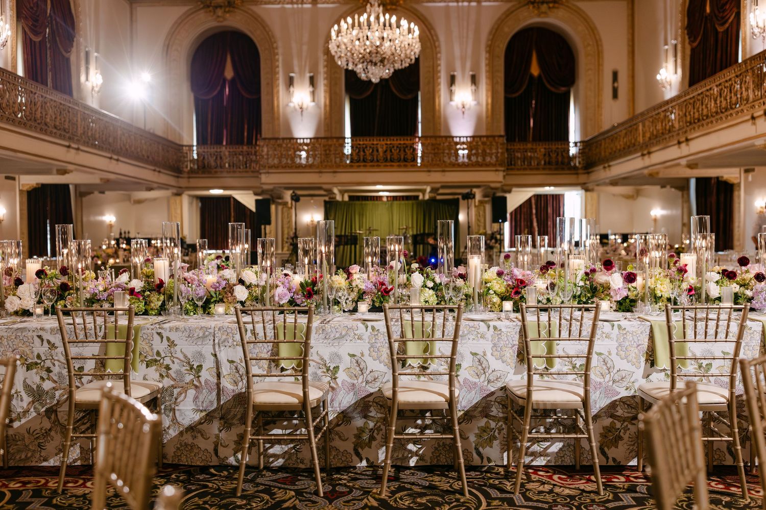 Elegant ballroom wedding reception with crystal chandeliers, white floral centerpieces, and gold chiavari chairs.