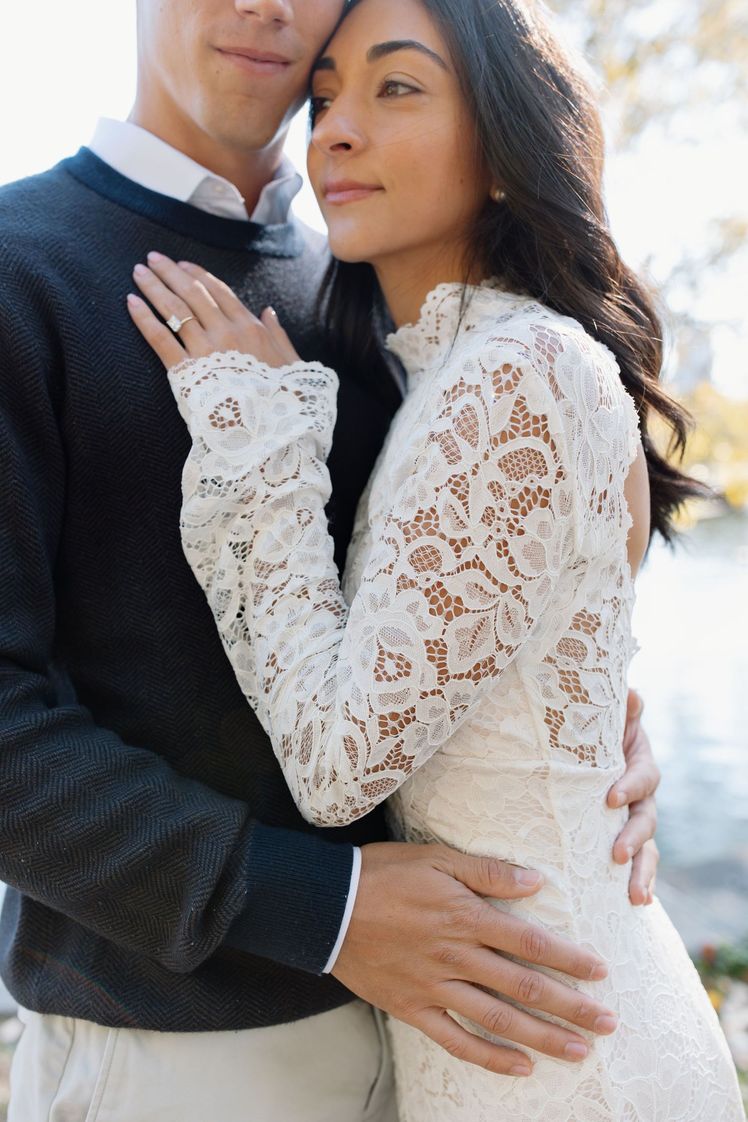 A romantic wedding couple embraces outdoors, with the bride wearing a delicate long-sleeve lace dress.