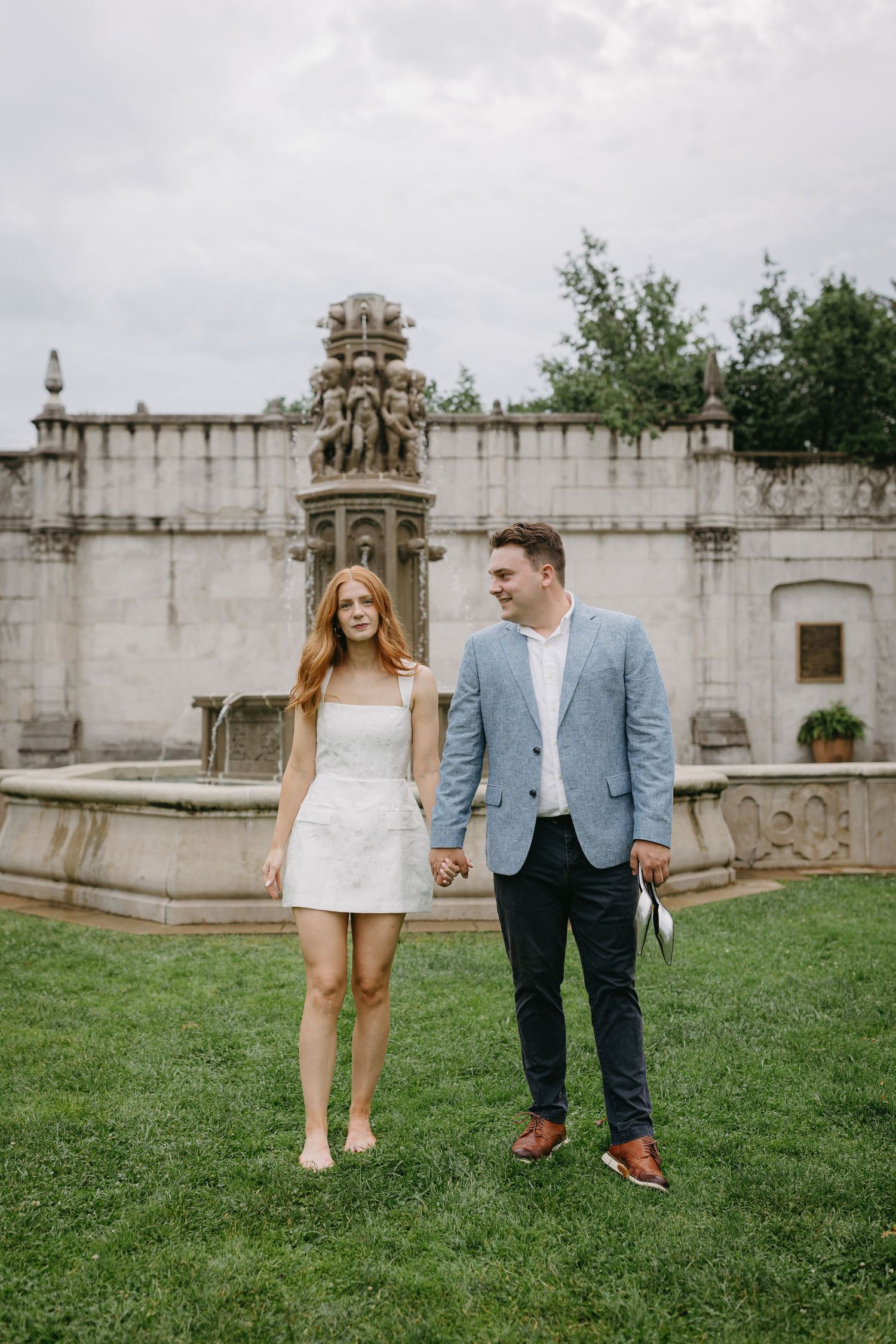 A couple in casual attire walking and posing playfully on grass near an ornate stone wall and architectural elements.