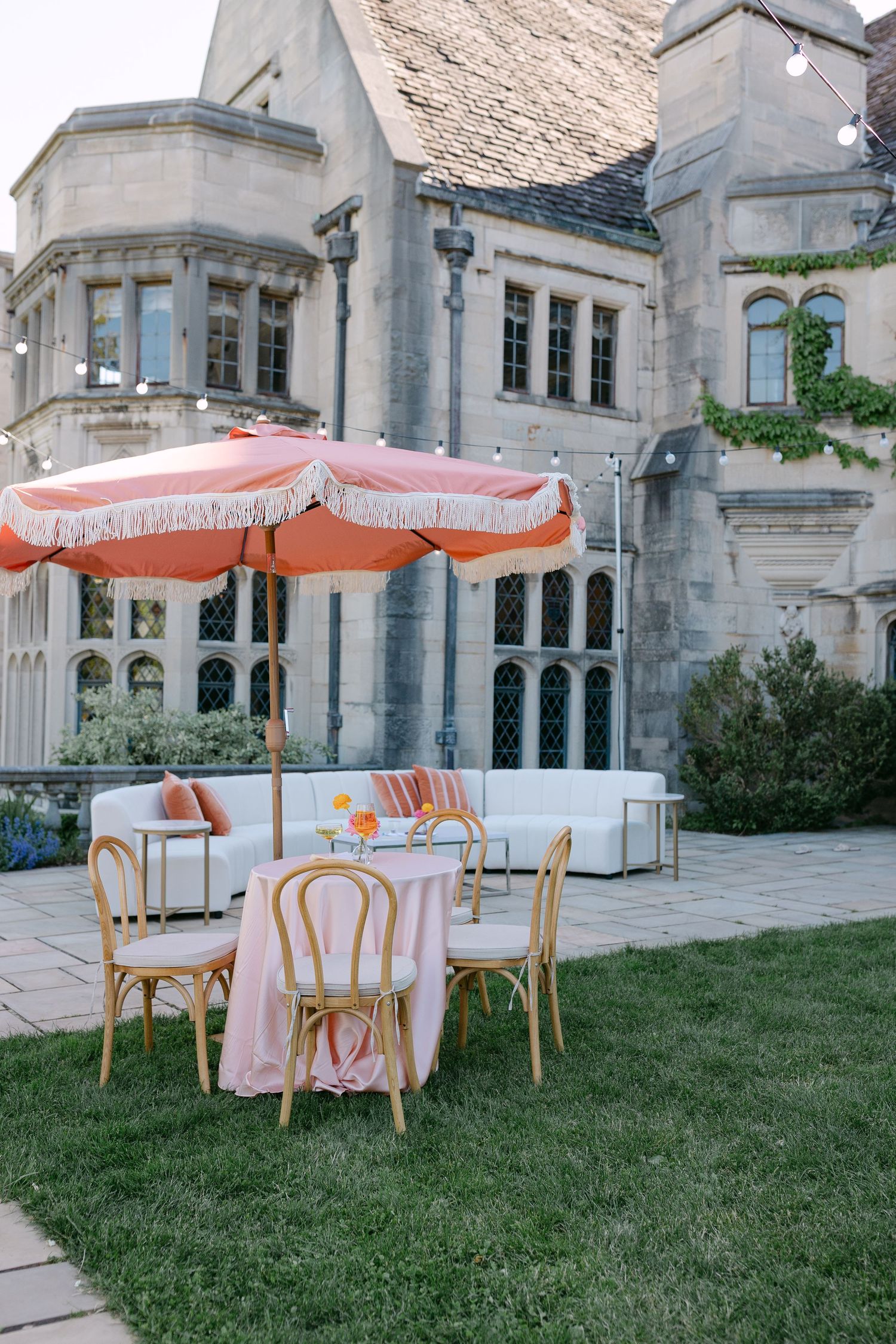 Elegant outdoor wedding setup with rattan chairs, pink tablecloth, and orange fringed umbrella on a stone mansion patio.