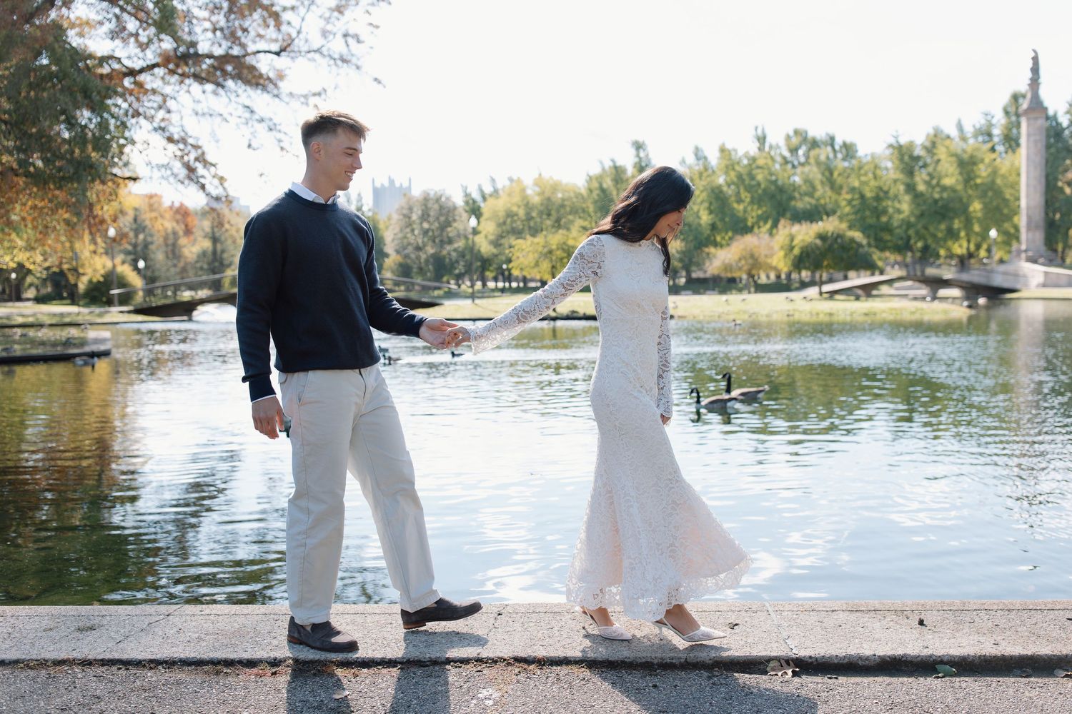 A couple holds hands while walking along a scenic lakeside path on a sunny autumn day.