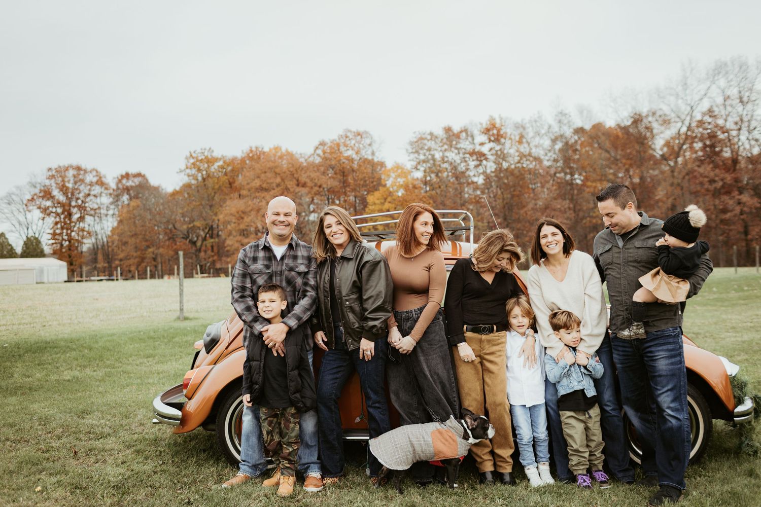 A group poses casually against a vintage car in an autumn field setting.