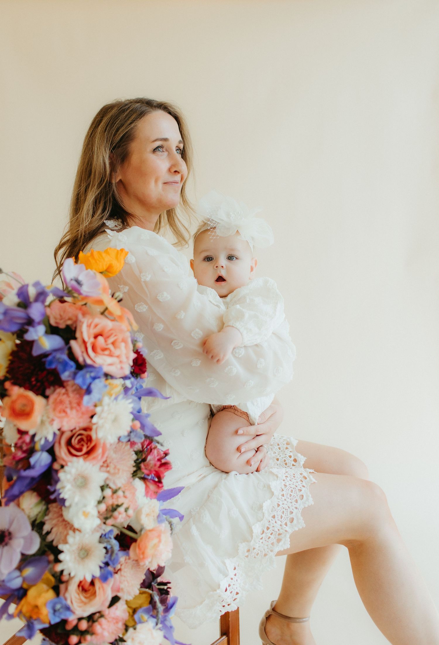 Mother and baby in matching white dresses pose with vibrant floral arrangement.