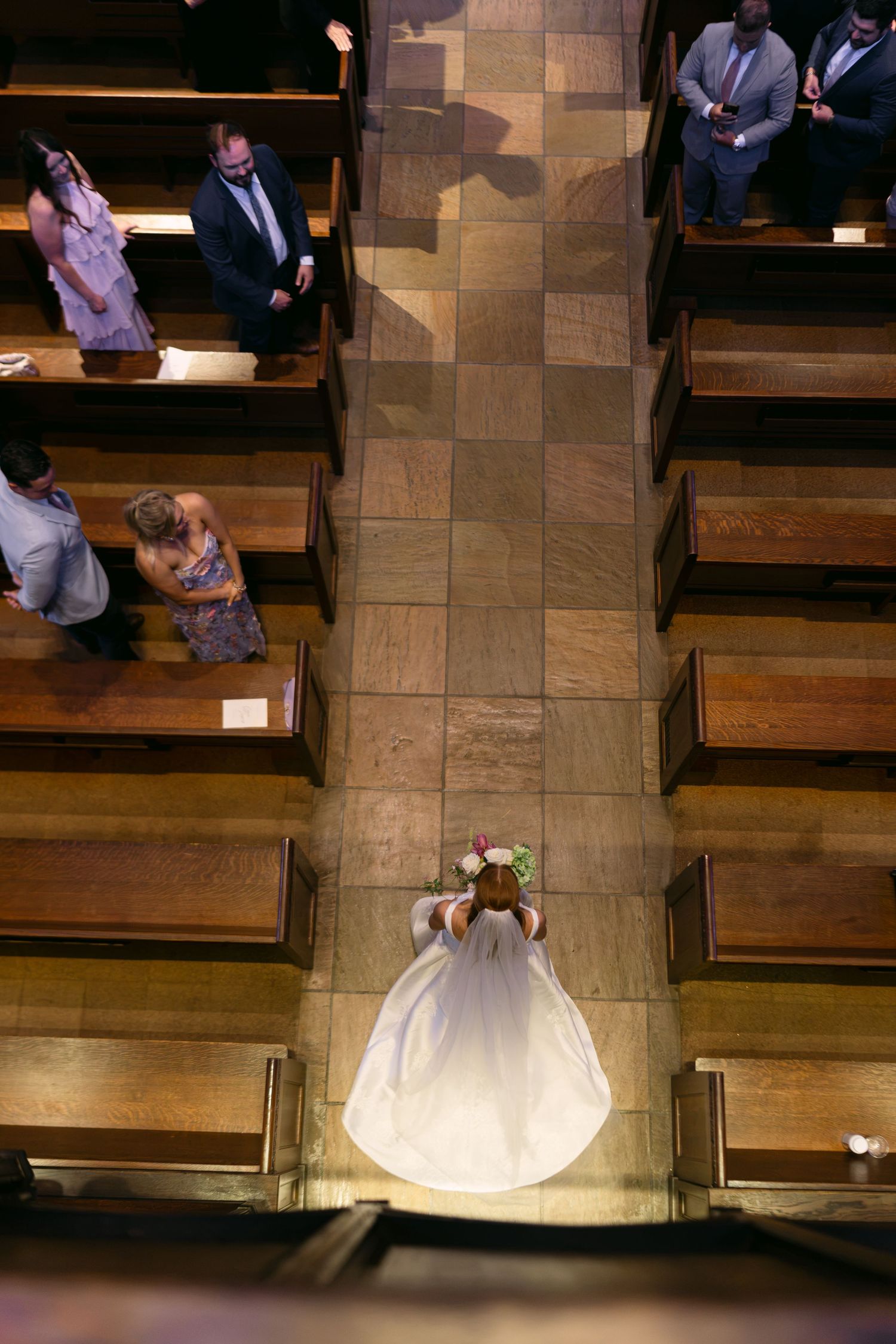 Aerial view of a bride in a white gown walking down the church aisle, with guests seated in wooden pews.