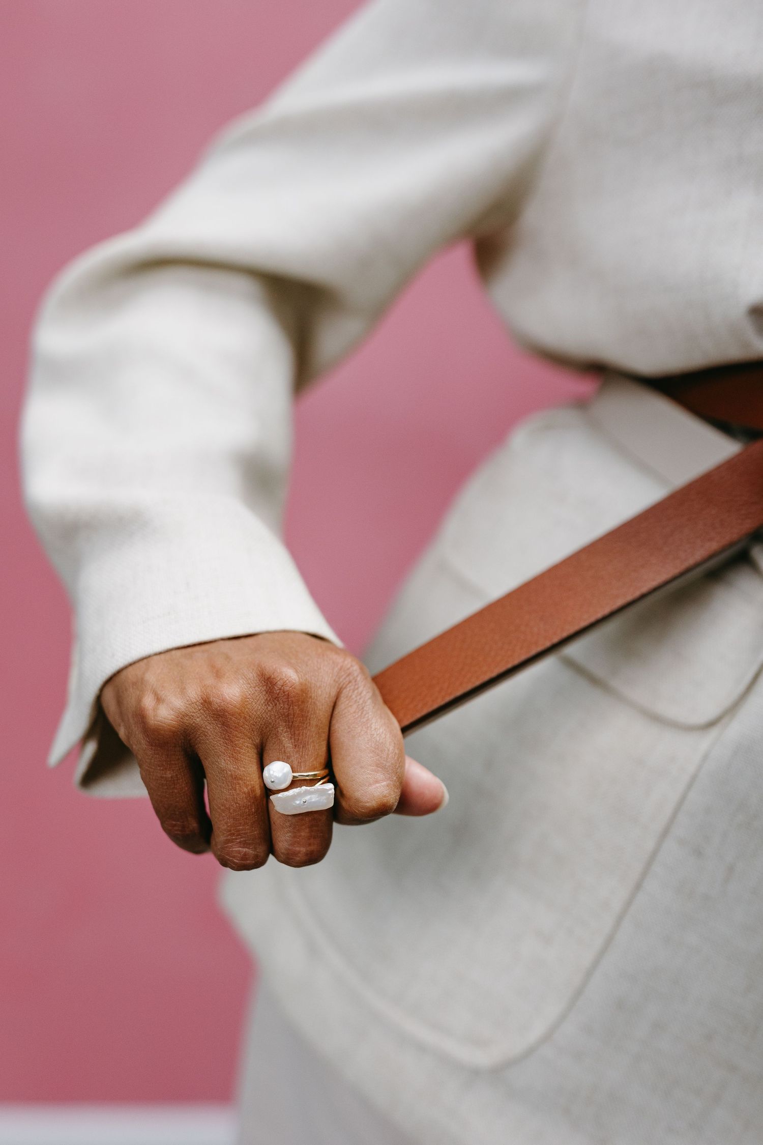 Close-up of hand wearing a silver ring while holding brown leather strap against white fabric and pink background.