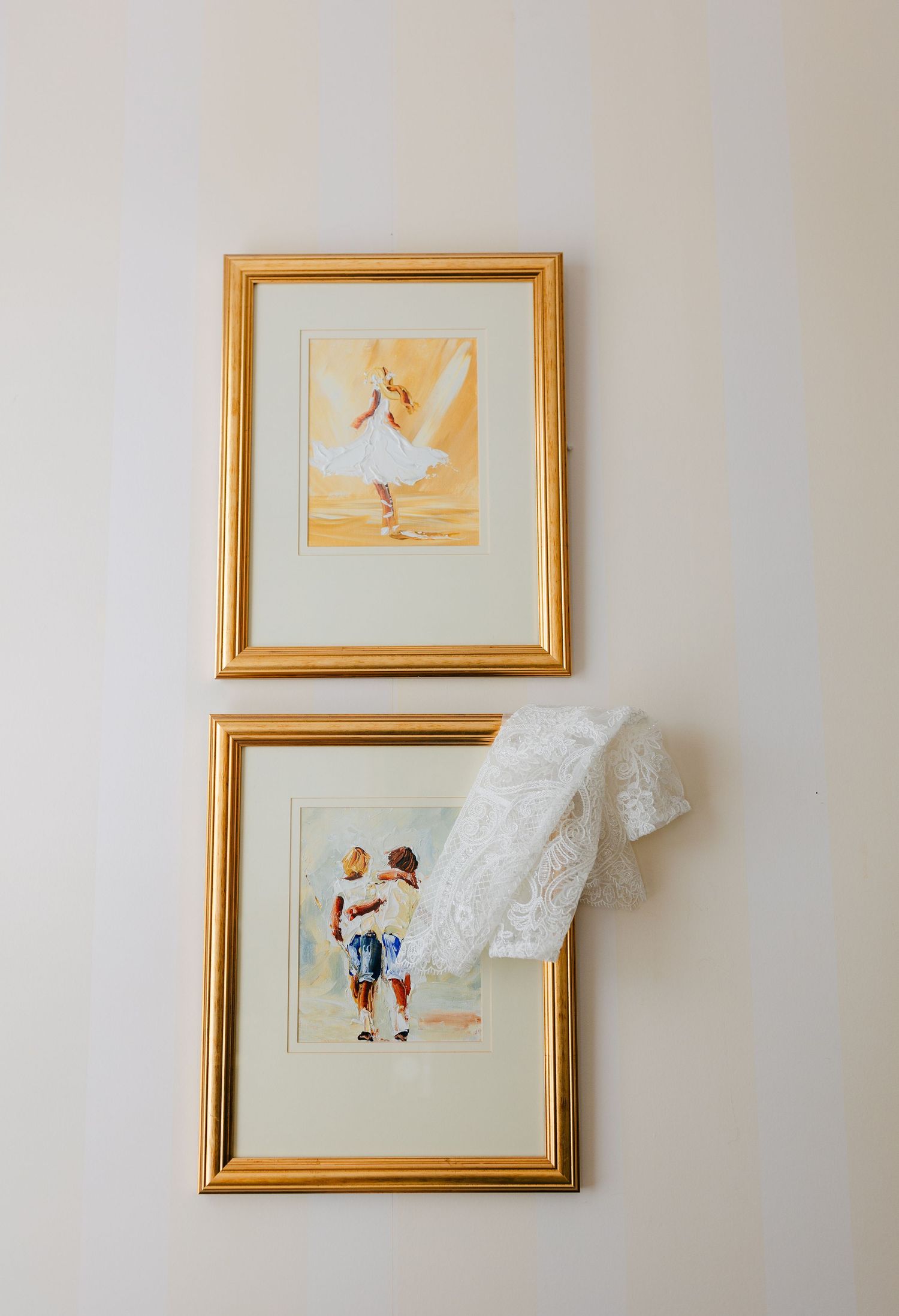 Two framed vintage-style artworks hang on a white wall with a piece of delicate white lace draped over the lower frame.