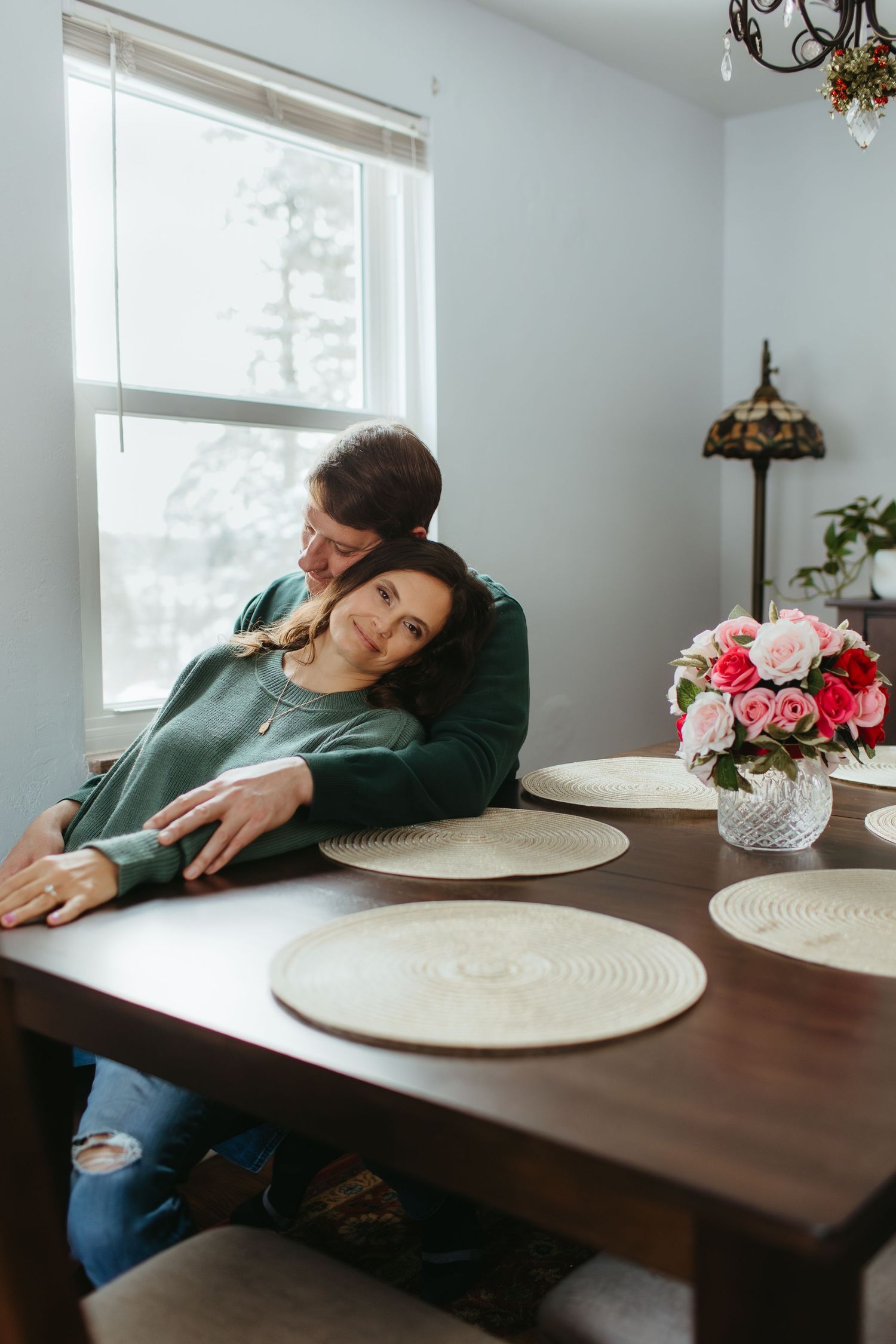 Couple relaxing at dining table with floral centerpiece near window.