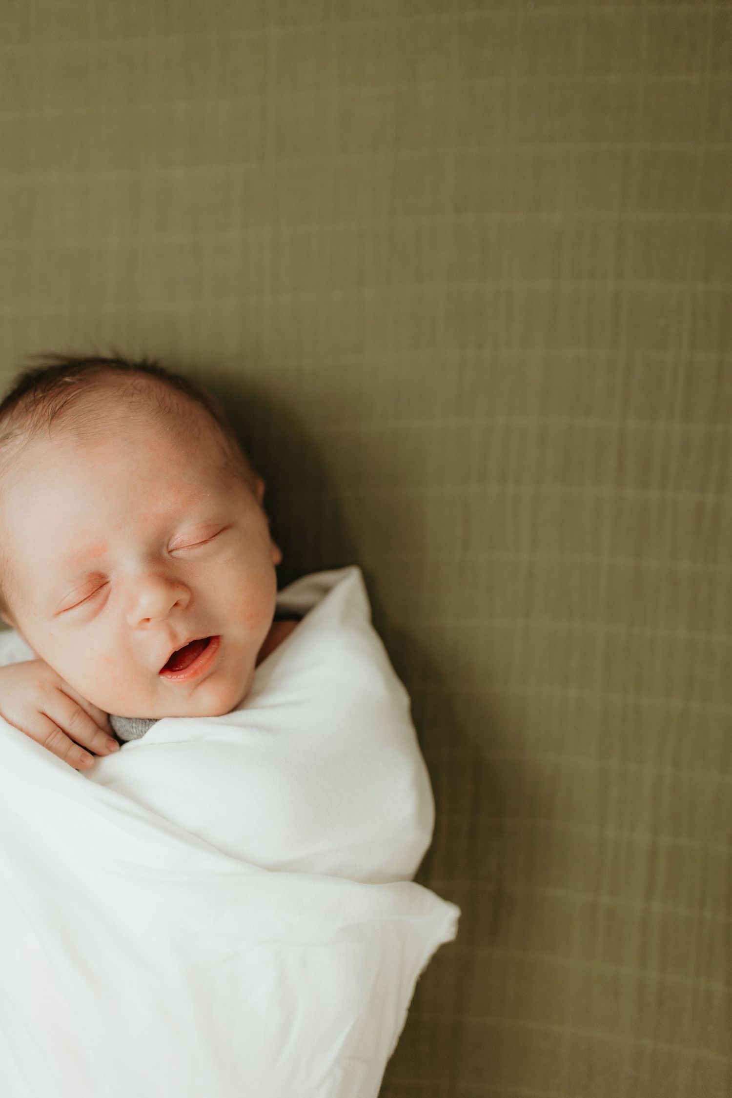 A peaceful newborn baby sleeping wrapped in a white blanket against a textured background.