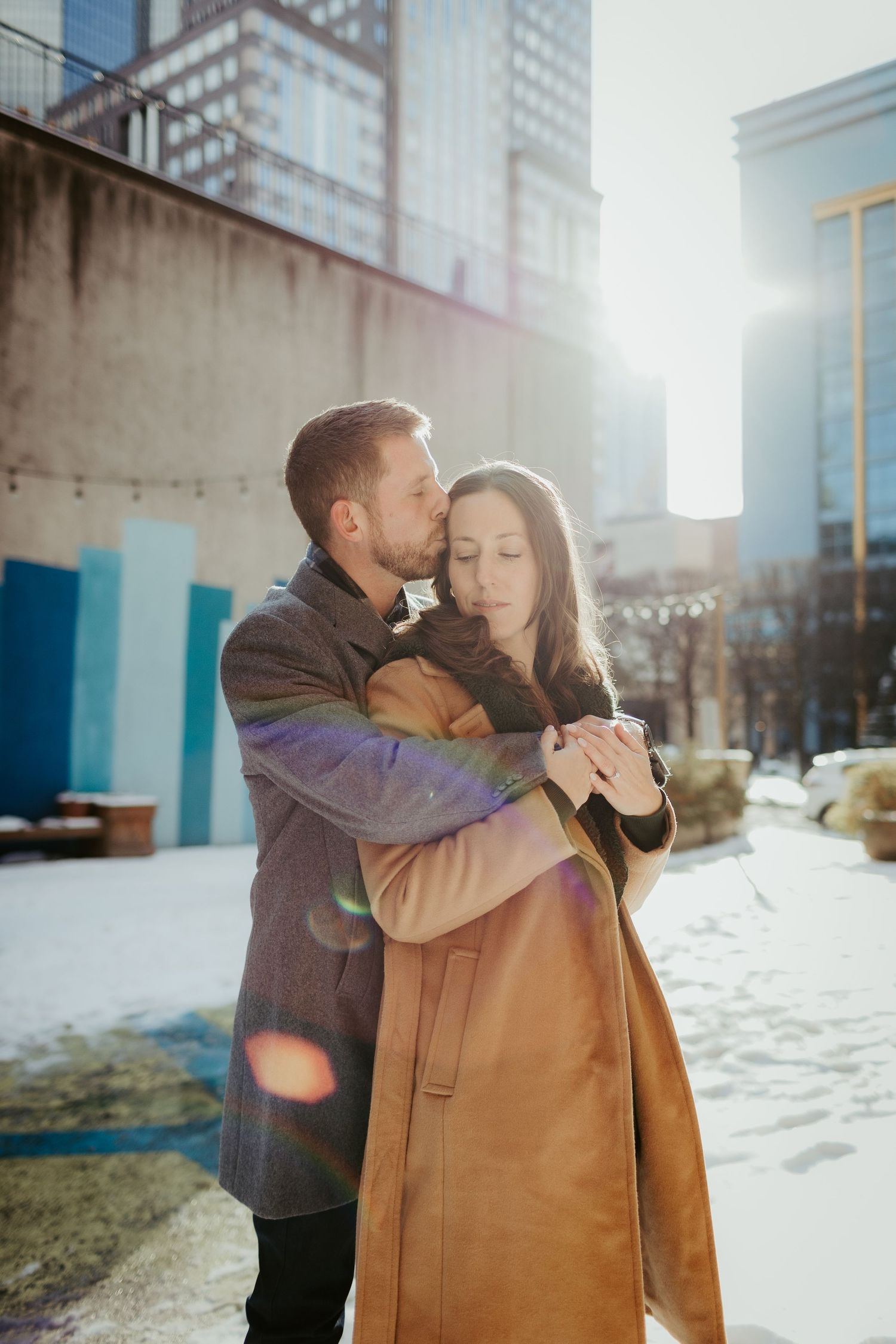 A couple shares an intimate moment in an urban setting with sunlit skyscrapers in the background.