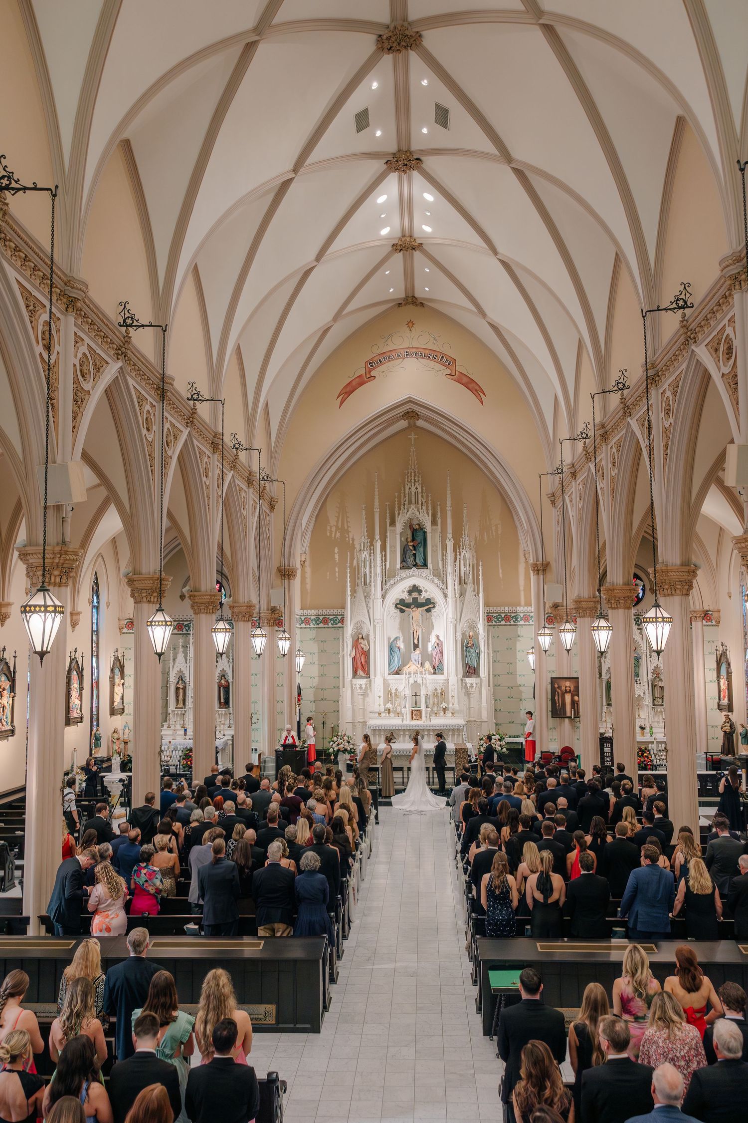 A crowded church ceremony with gothic vaulted ceilings and white arches filled with people in formal attire.