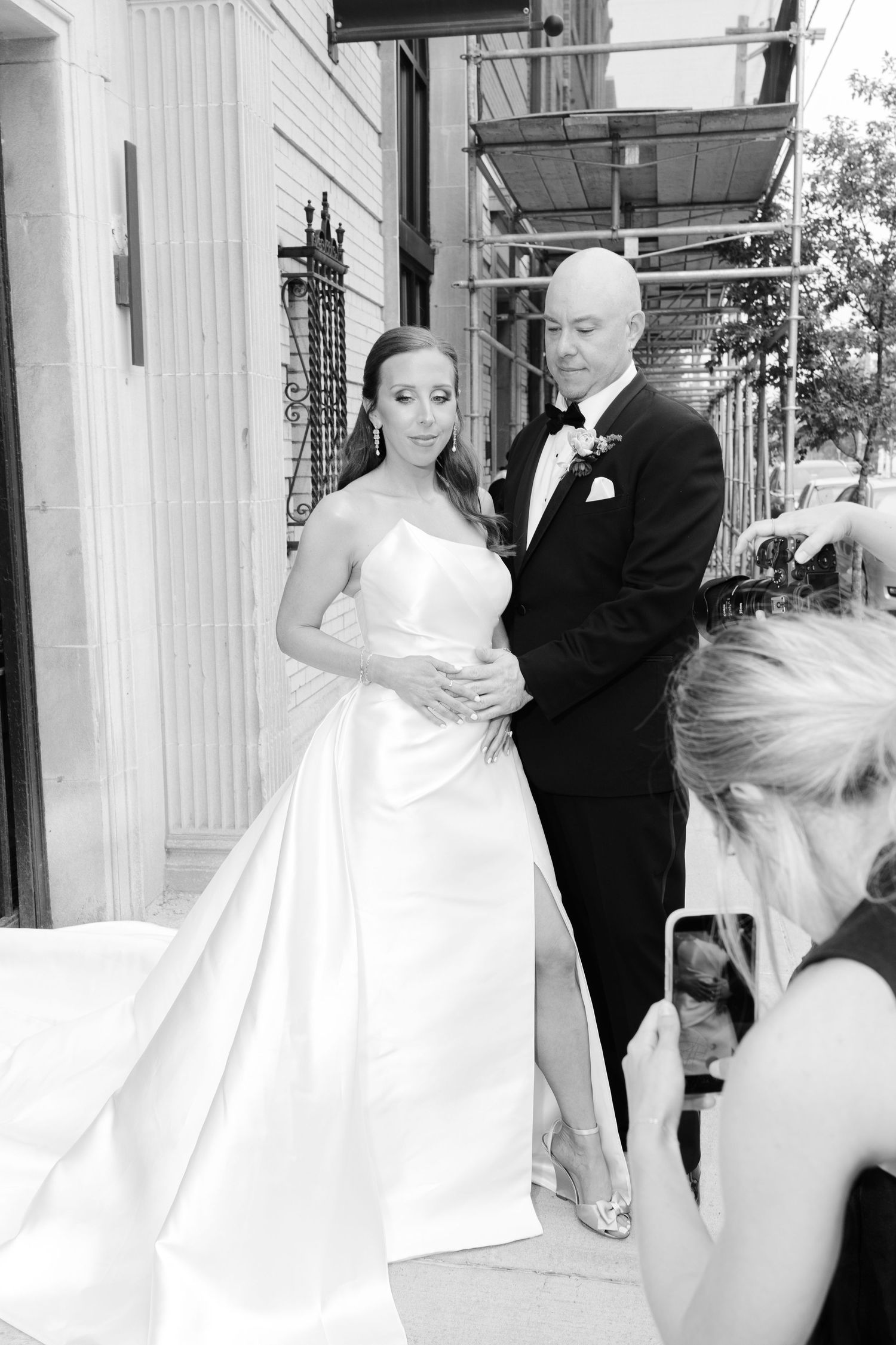 Black and white photo of a bride in a strapless gown and groom in a tuxedo posing outside while a guest photographs them.