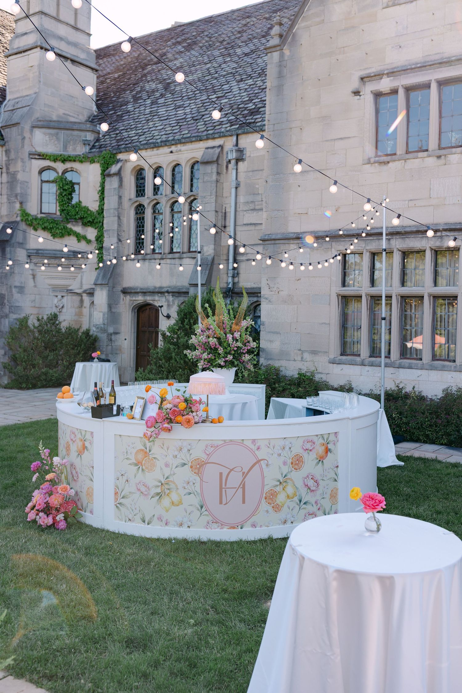 Outdoor wedding bar with floral design and monogram, string lights, and stone mansion backdrop at dusk.