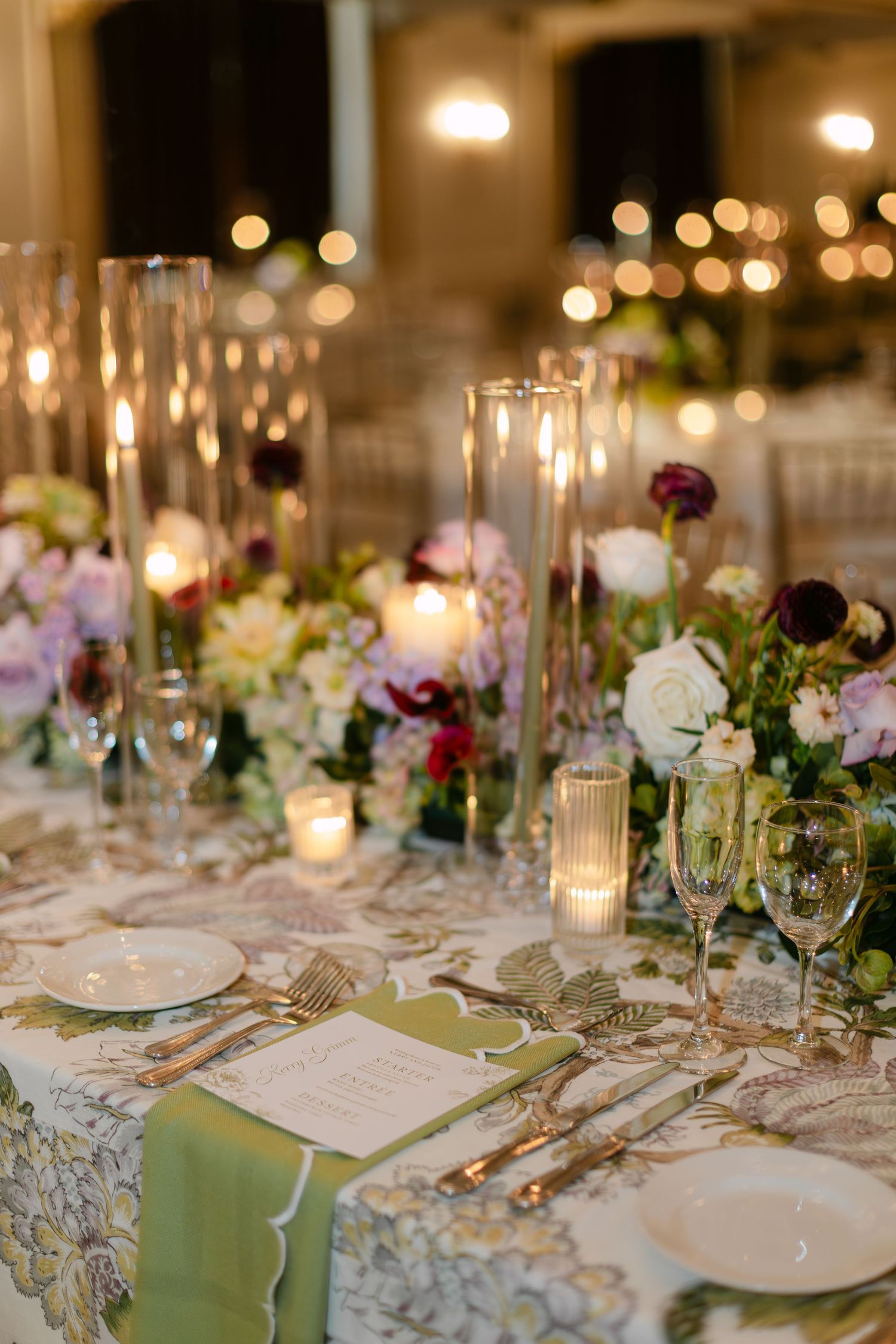 Elegant wedding reception table setting with candlelight, crystal glassware, and purple and white floral centerpieces.