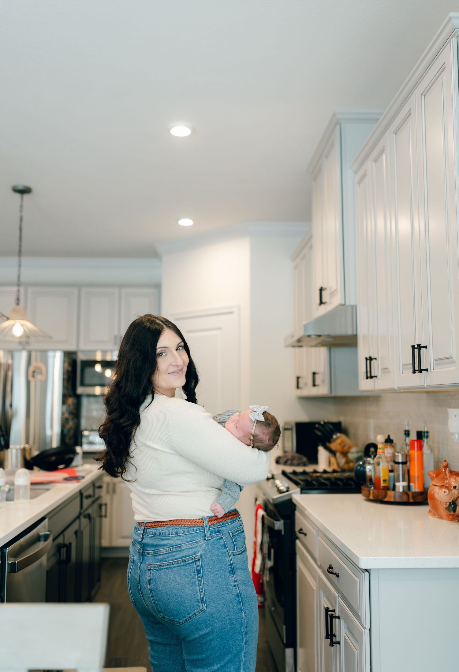 A casual home photo of a mother holding her baby while standing in a modern white kitchen.