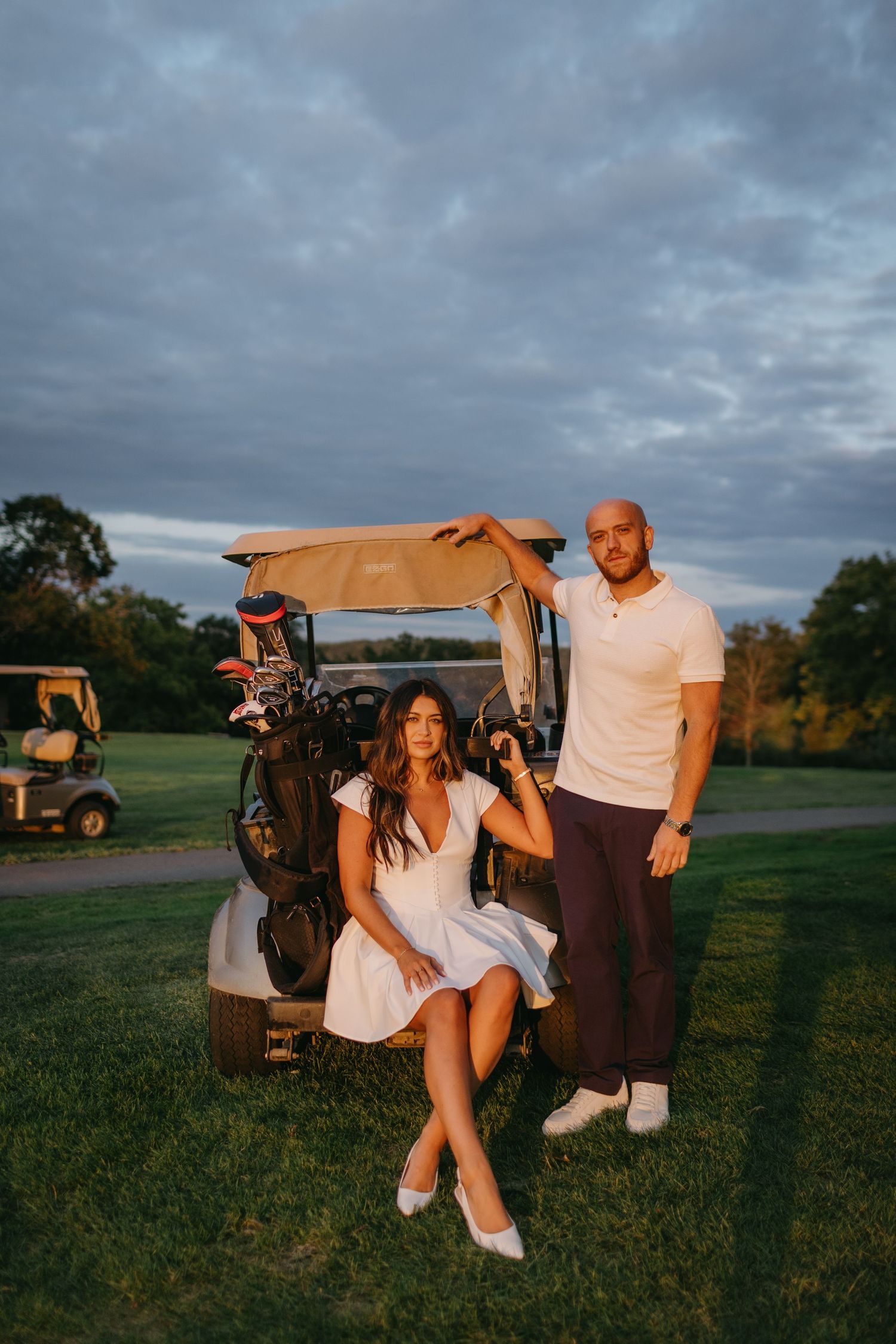 Casual couple poses with golf cart on grassy course during golden hour.