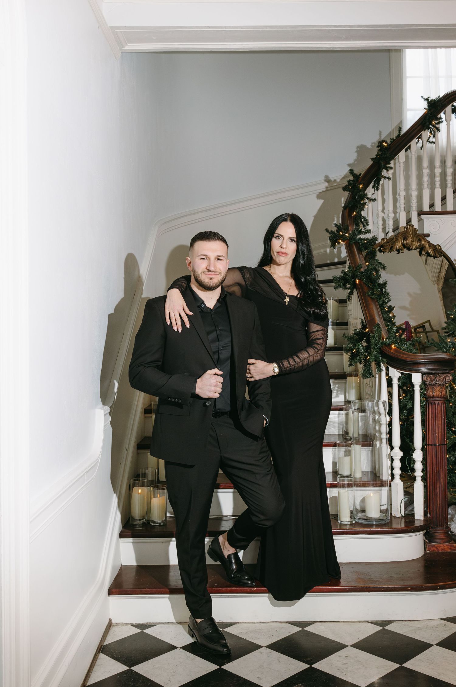 Couple in formal black attire posing on elegant staircase with holiday greenery.