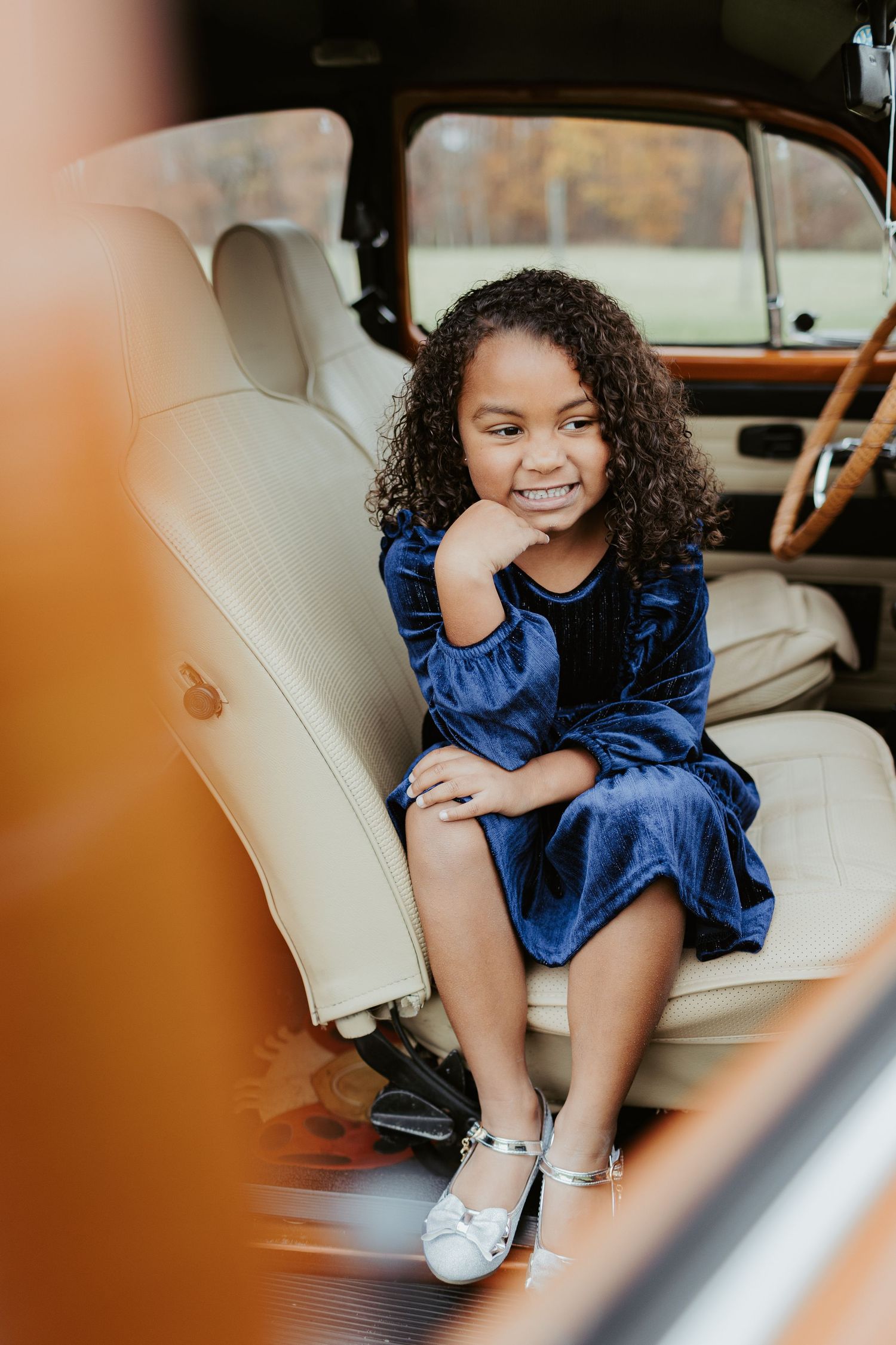 A child in a blue dress sits smiling on cream leather seats inside a classic car.