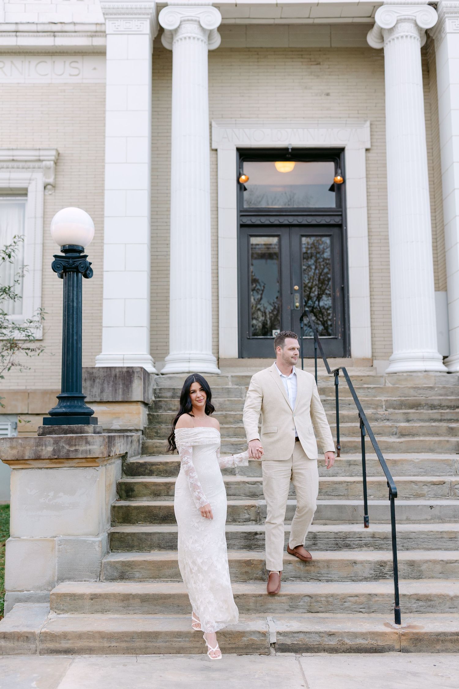 A couple in light-colored formal attire poses on the steps of a historic building with white columns.