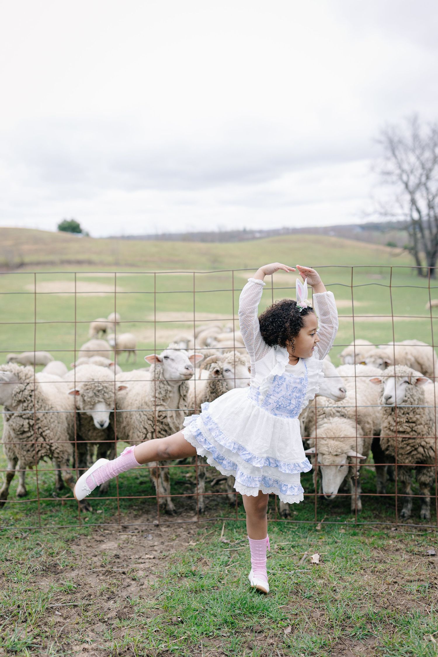 A ballerina in white tutu and leggings dances gracefully in a field beside a flock of sheep.