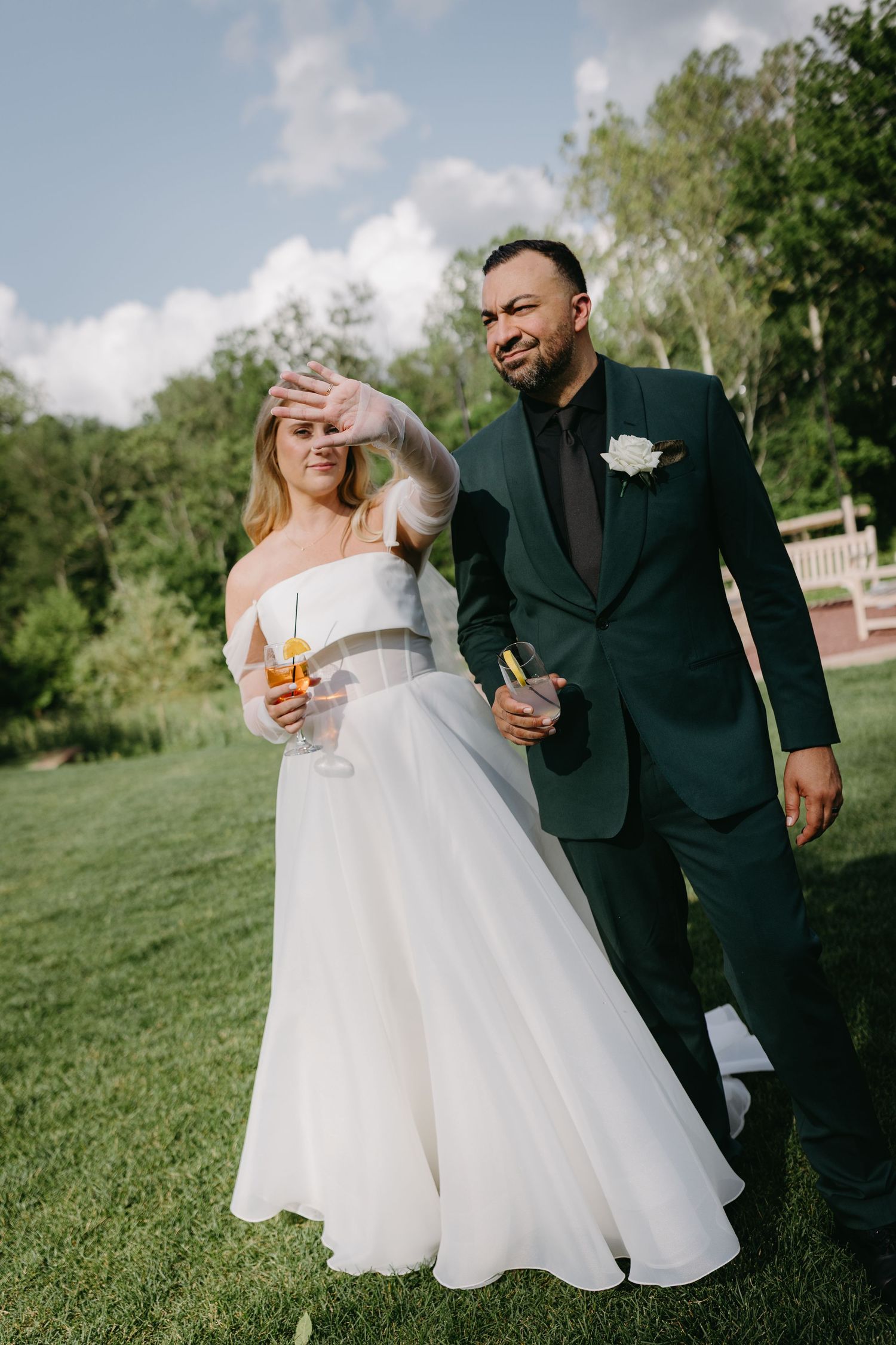 Wedding couple laughs together on a grassy lawn with trees in the background while holding drinks.