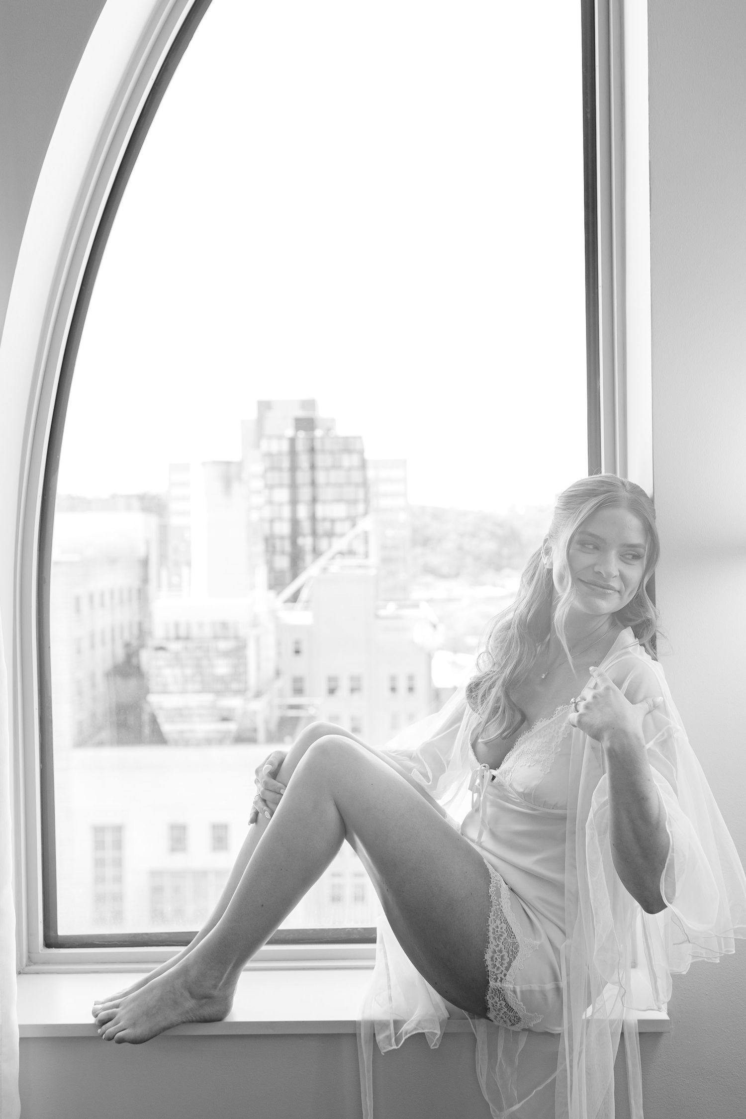Woman in white lace lingerie sitting on a window sill with city skyline in black and white photo.