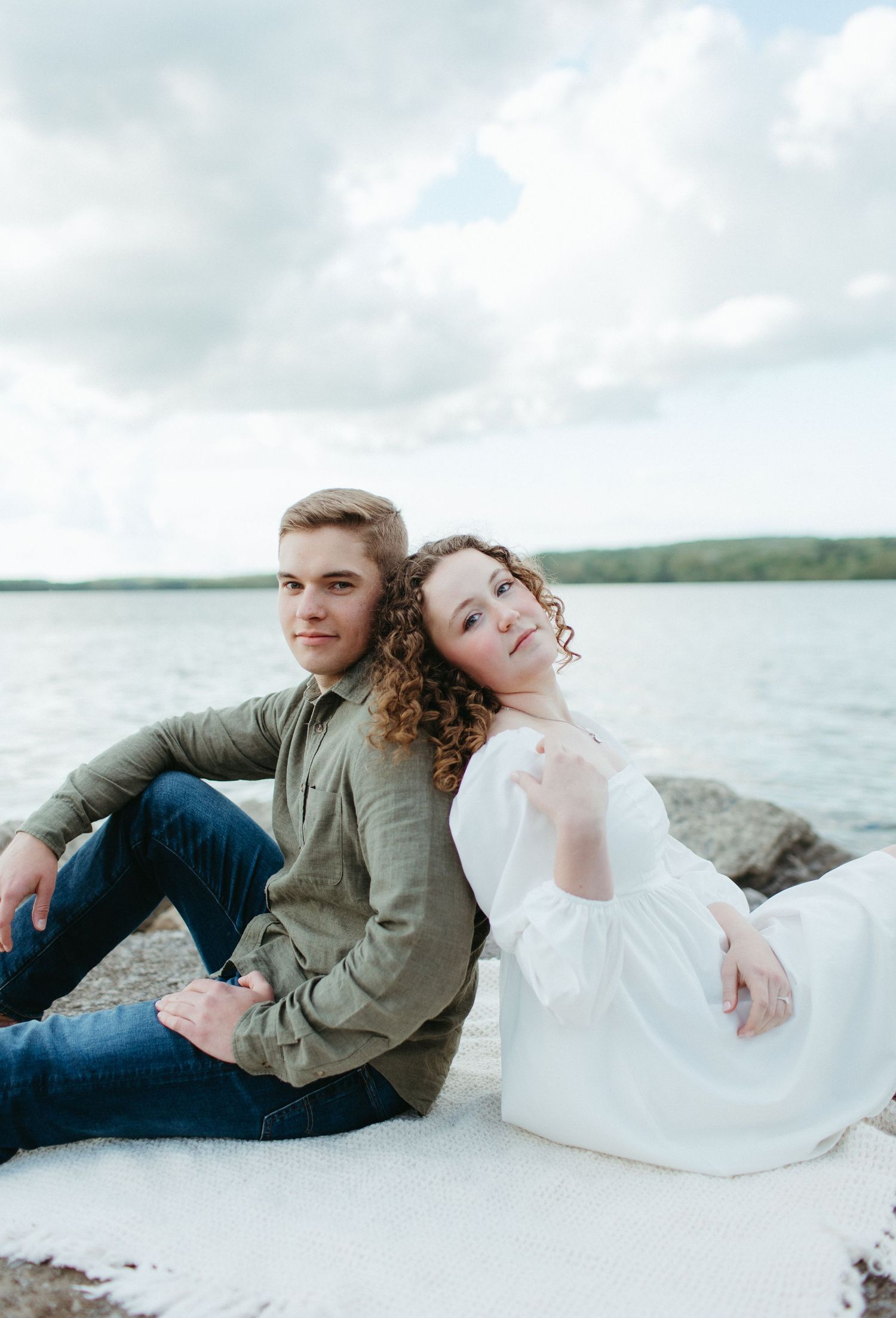 Couple sitting back to back on rocky shore with lake and cloudy sky in background.
