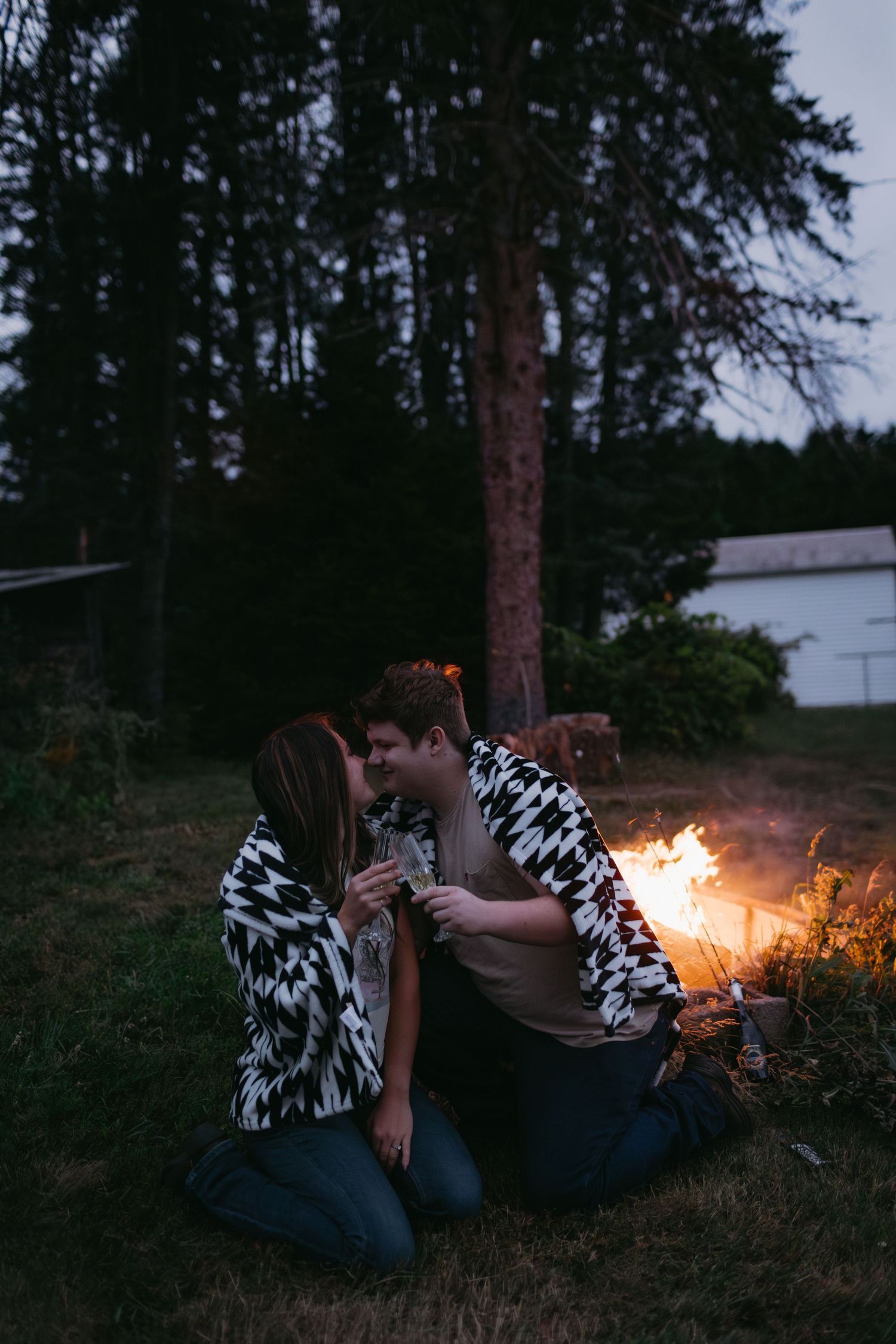 Couple in matching blankets sharing intimate moment by campfire at dusk.