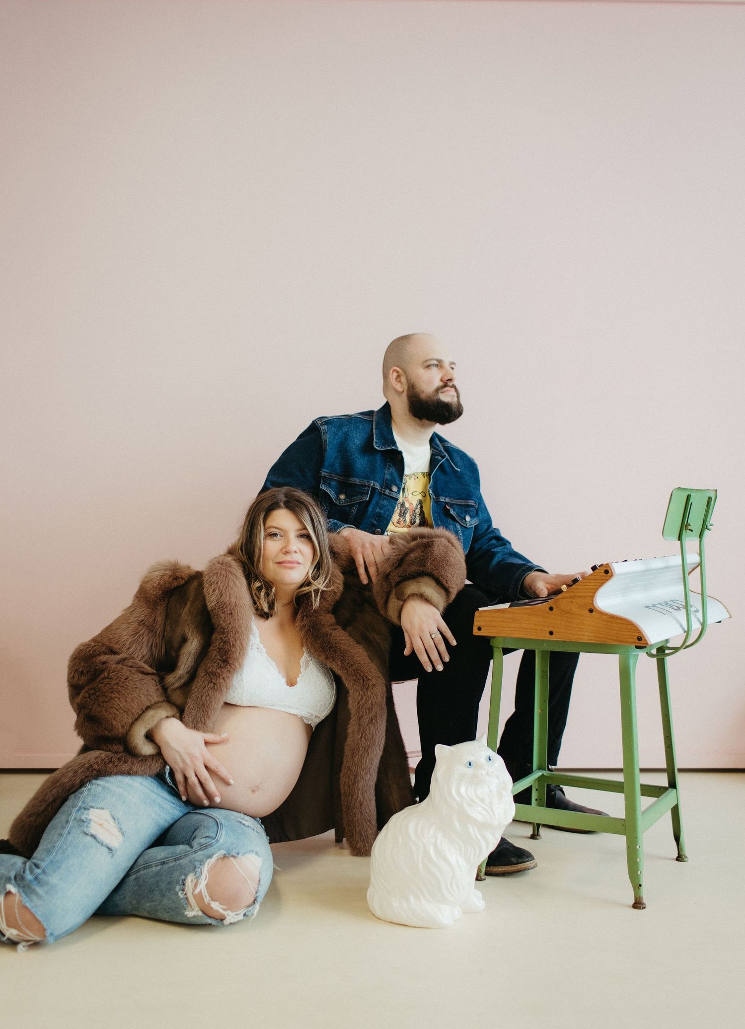 Studio maternity photo with couple and green chair against pink background.