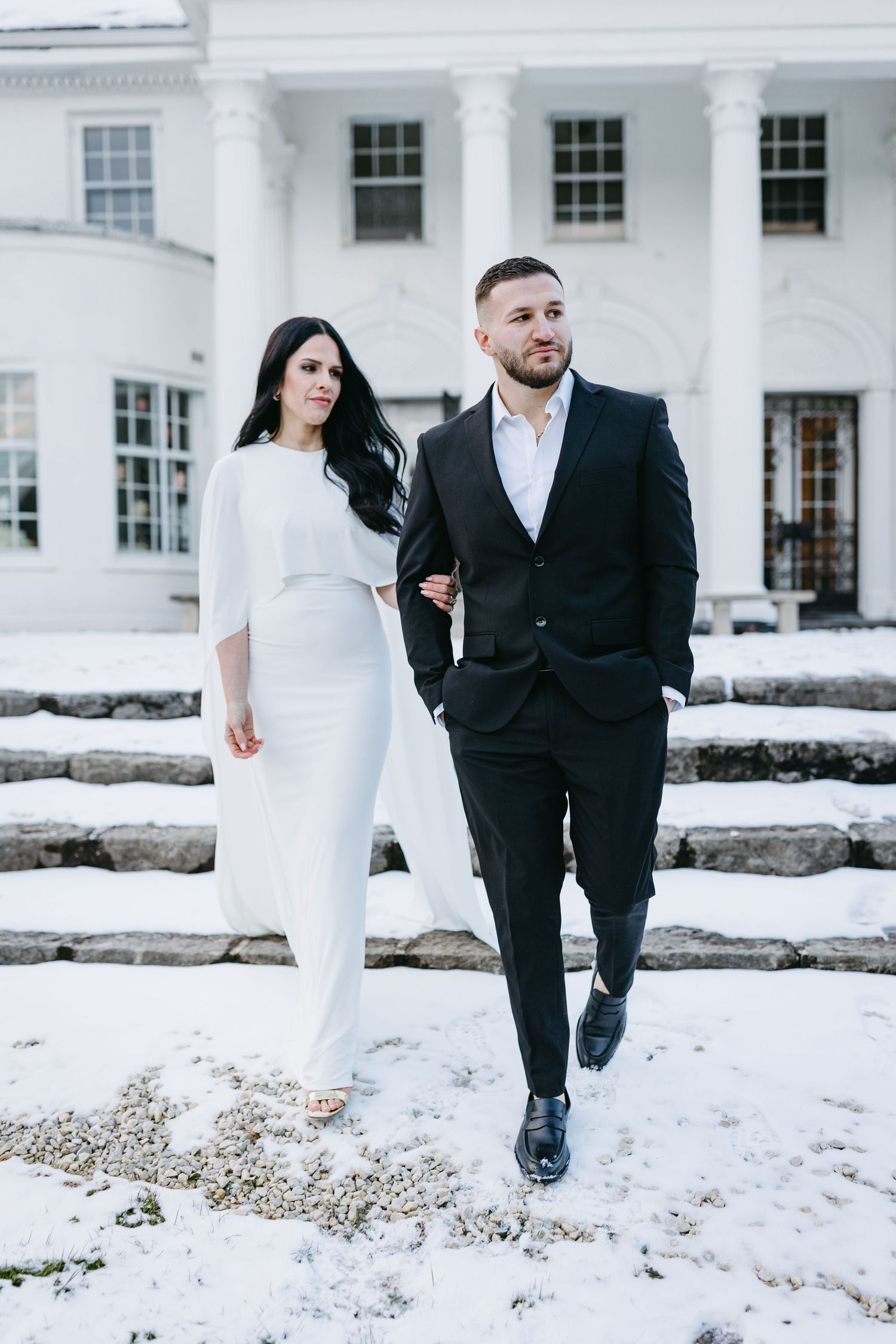 Couple in formal wear walking down snowy steps of white columned building.