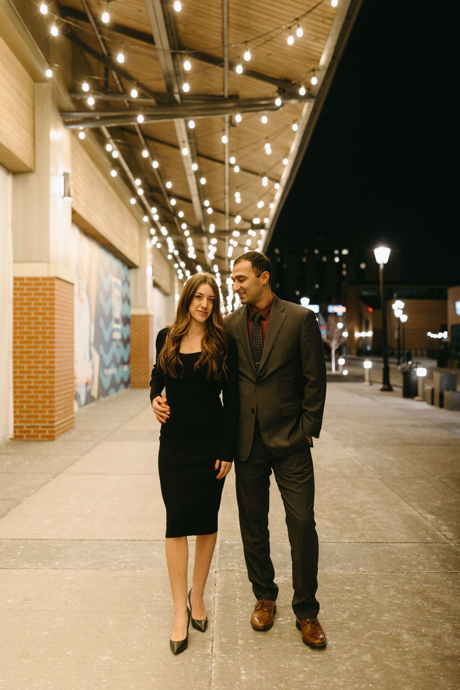 Couple walking together under romantic string lights at night along urban pathway.