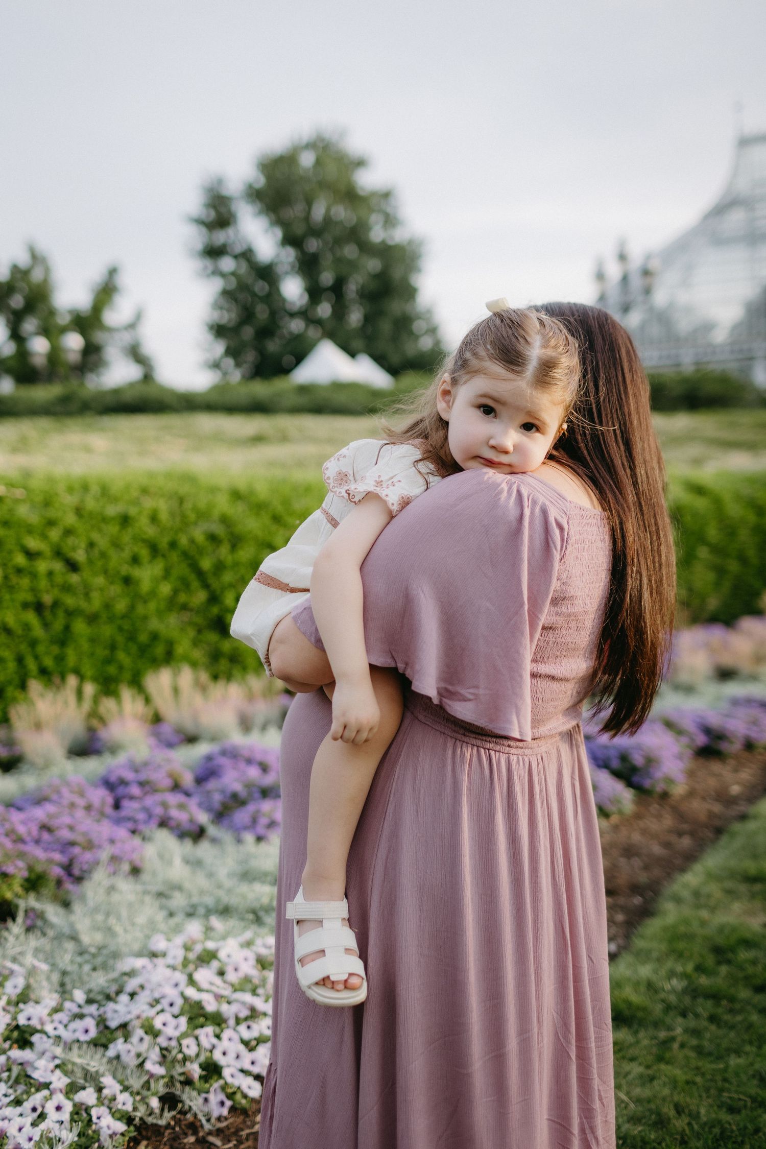A tender moment in a garden as a figure in a dusty rose dress holds a small child near blooming purple flowers.